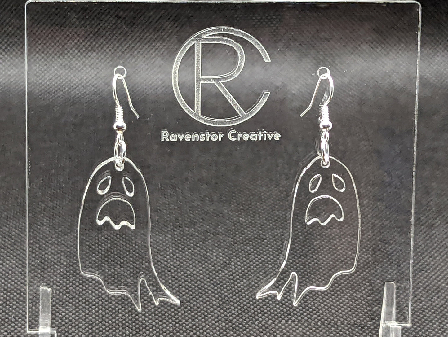 Shows a pair of earrings with silver ear wire hook fixings. They are spooky moaning ghost shapes. The colour of them is clear/see through. They are on a see through stand that has the Ravenstor Creative Logo and its against a dark background.