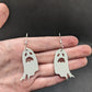 Image of a pair of spooky moaning Ghost shaped earrings on a hand showing the size against a dark background, they are two and a half finger widths length  and two wide. They are grey and have silver fixings.