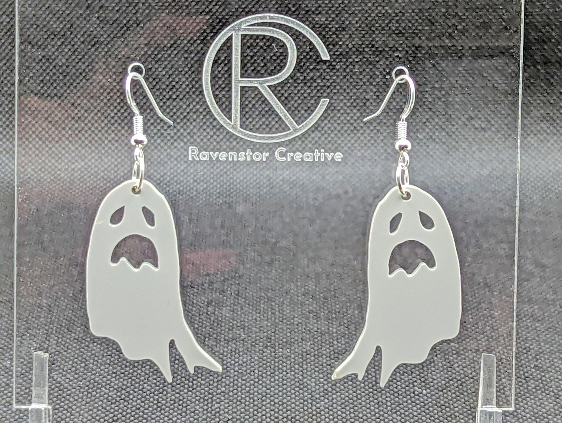 Shows a pair of earrings with silver ear wire hook fixings. They are spooky moaning ghost shapes. The colour of them is grey. They are on a see through stand that has the Ravenstor Creative Logo and its against a dark background.