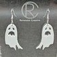 Shows a pair of earrings with silver ear wire hook fixings. They are spooky moaning ghost shapes. The colour of them is white. They are on a see through stand that has the Ravenstor Creative Logo and its against a dark background.