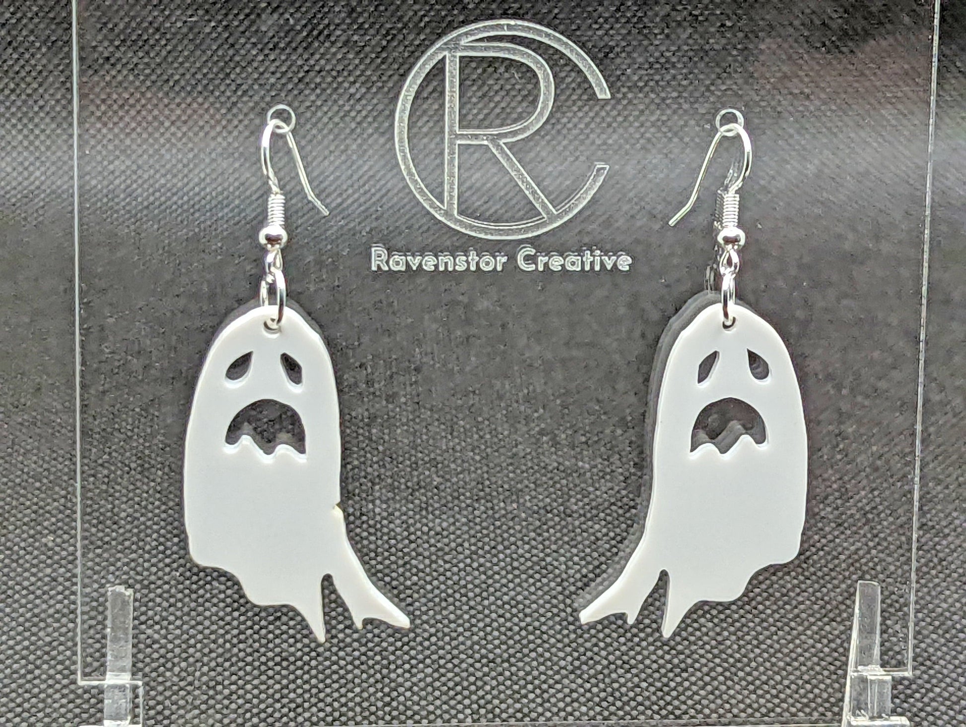 Shows a pair of earrings with silver ear wire hook fixings. They are spooky moaning ghost shapes. The colour of them is white. They are on a see through stand that has the Ravenstor Creative Logo and its against a dark background.