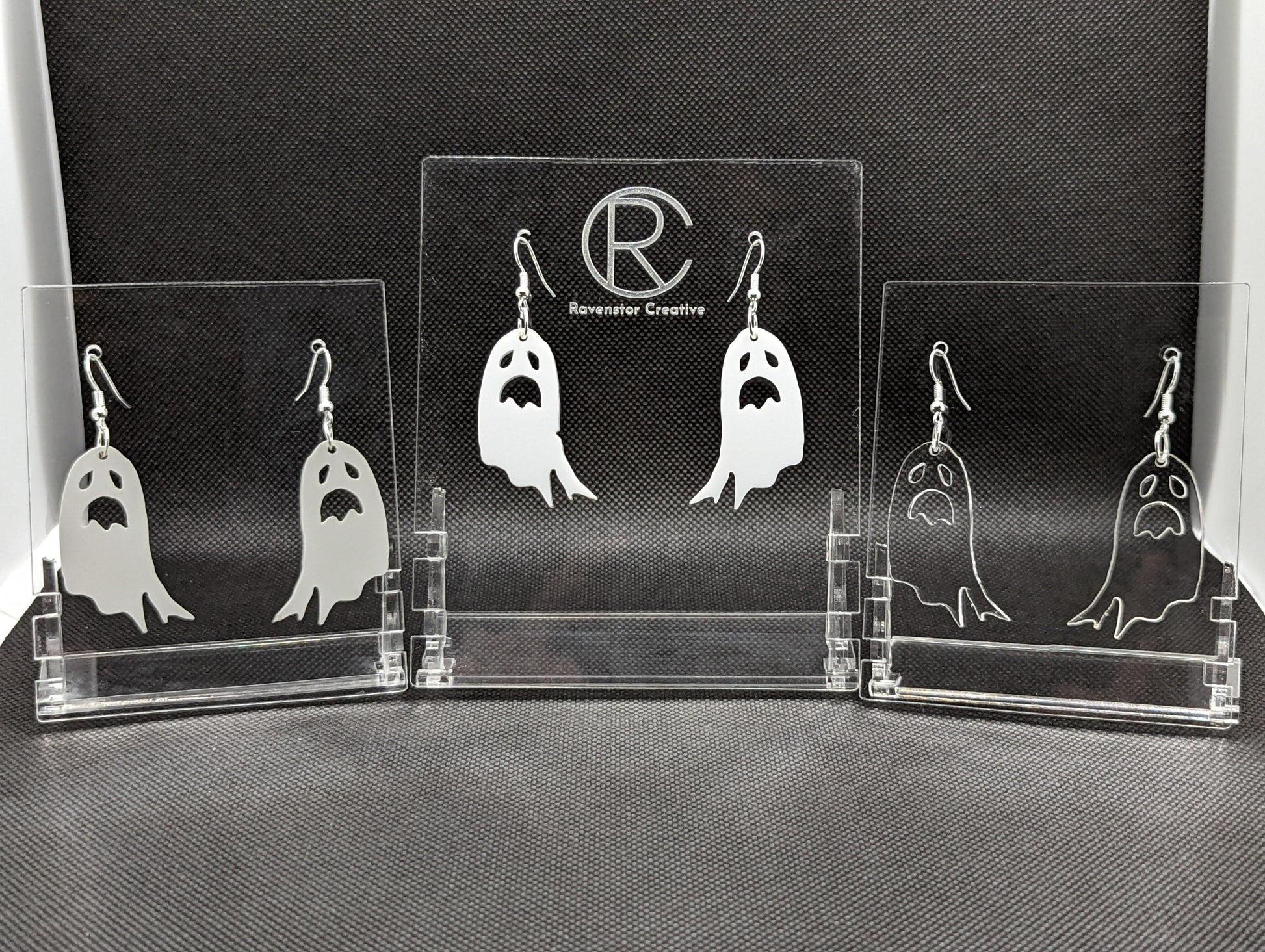 Shows 3 pairs of earrings with silver ear wire hook fixings. They are spooky moaning ghost shapes. The colour of them is Grey, White and clear/see through. They are on see through stands against a dark background.