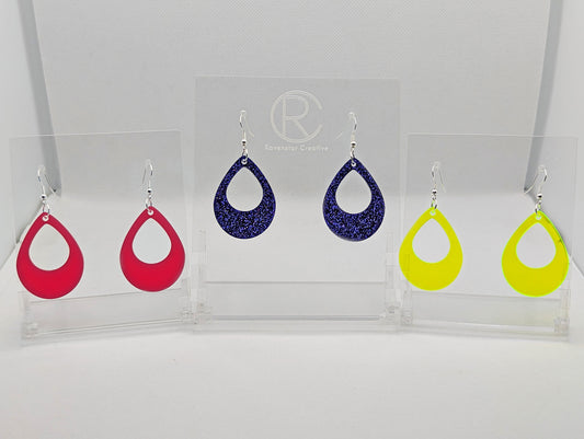 Shows three pairs of earrings that have ear wire hook fixings. They are teardrop shaped hoops. The colours are hot pink, purple glitter and neon orange. They are against a white background on see through stand with the ravenstor creative logo on.