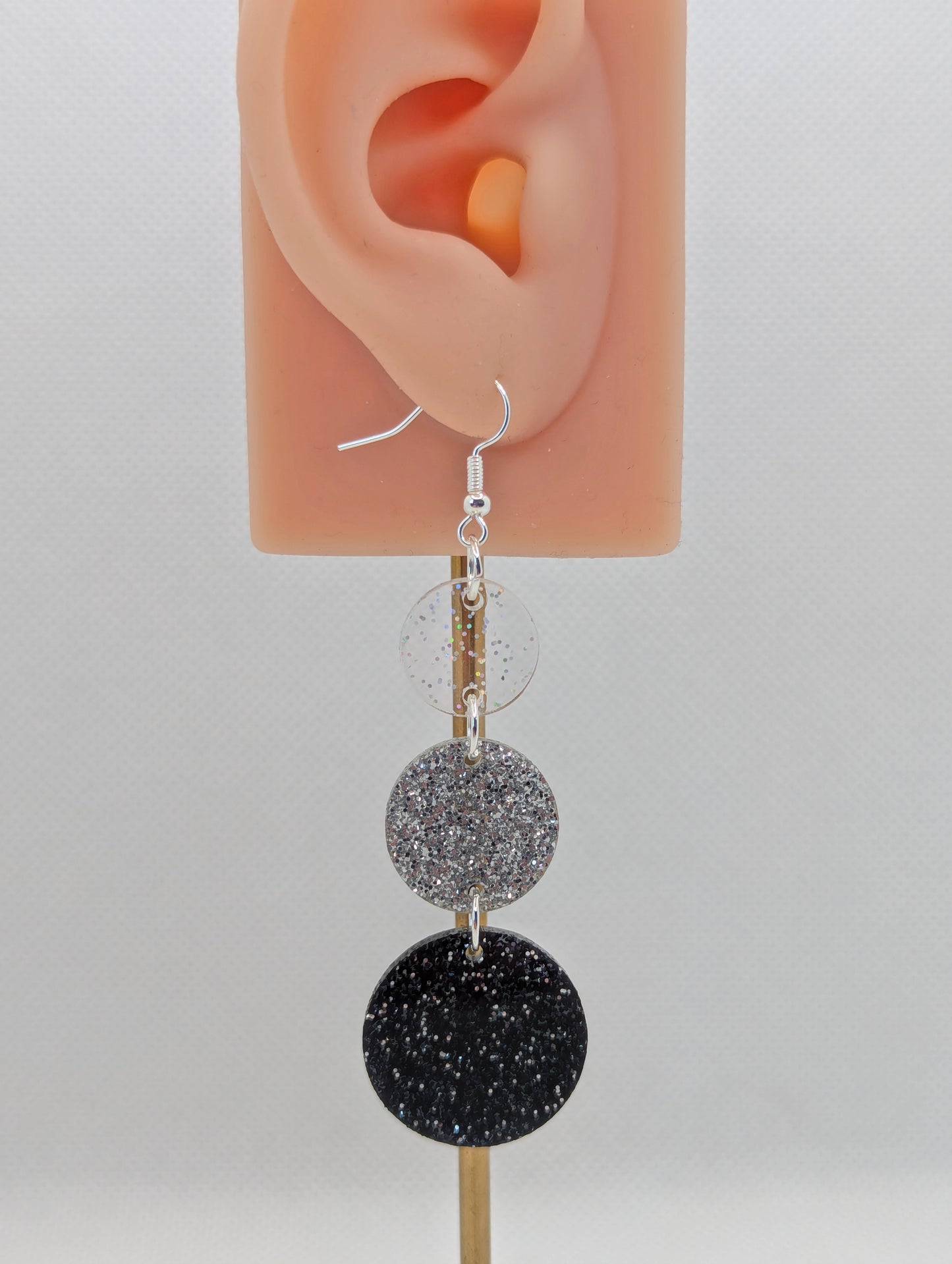 An earrings which has three circles dangling from each other, the top is the smallest and they get bigger as they go down. It is on an ear to show the size. It is made from Clear Sparkle, Silver and Black Glitter acrylics and have silver ear wire hook fixings.