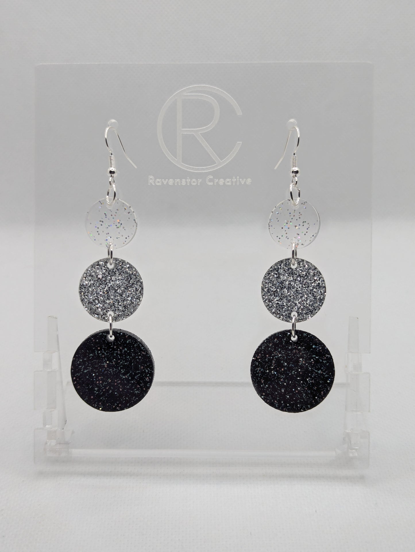 A pair of acrylic earrings with Silver ear wire hook fixings. Each has three circles dangling from each other, the top is the smallest and they get bigger as they go down. The colours from the top are Clear Sparkle, Silver Glitter and Black Glitter.