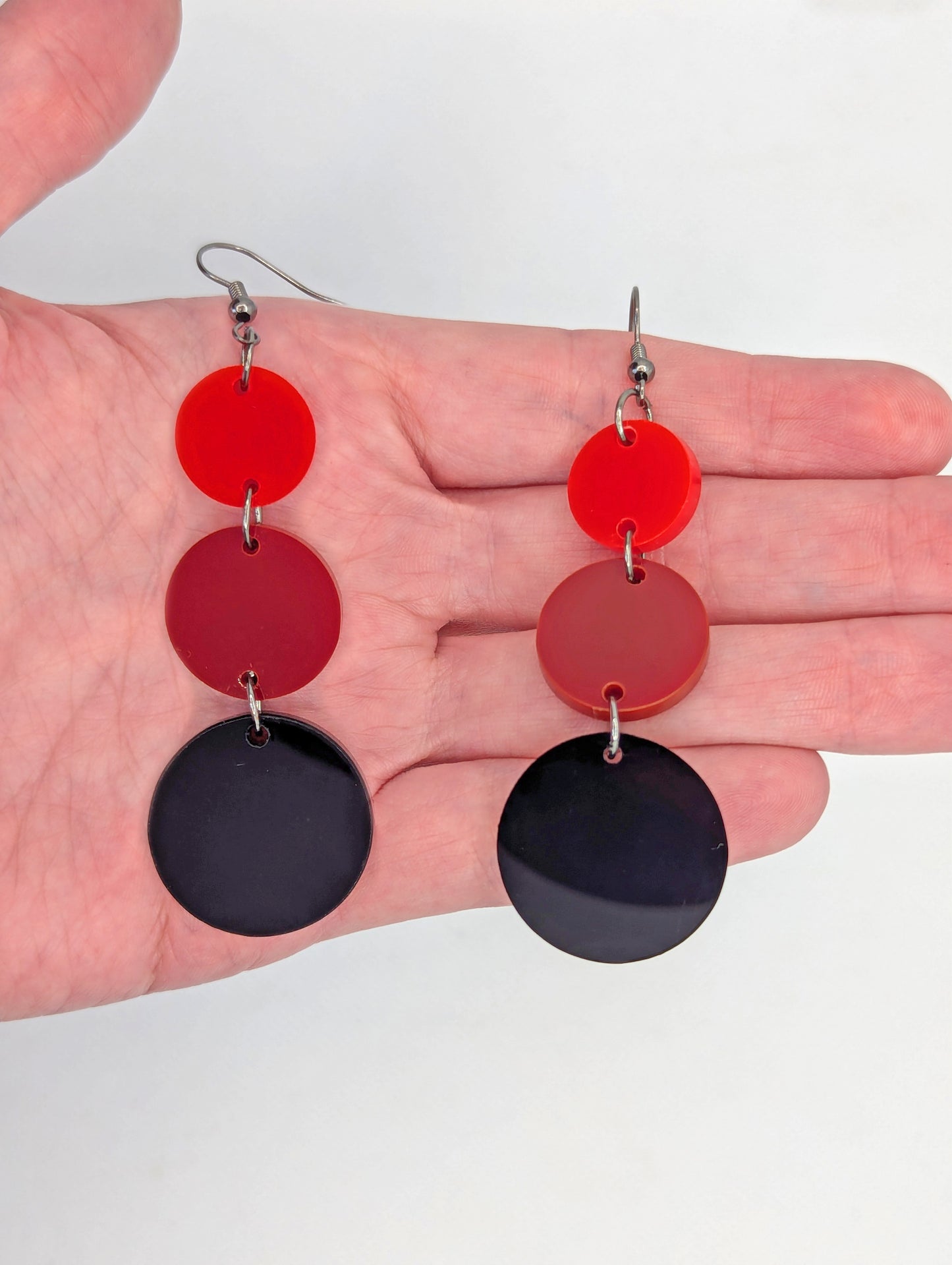 A pair of dangly earrings, each has three circles dangling from each other, the top is the smallest and they get bigger as they go down. They are against a hand to show the size. They are four finger widths in length and the bigger heart is two wide.