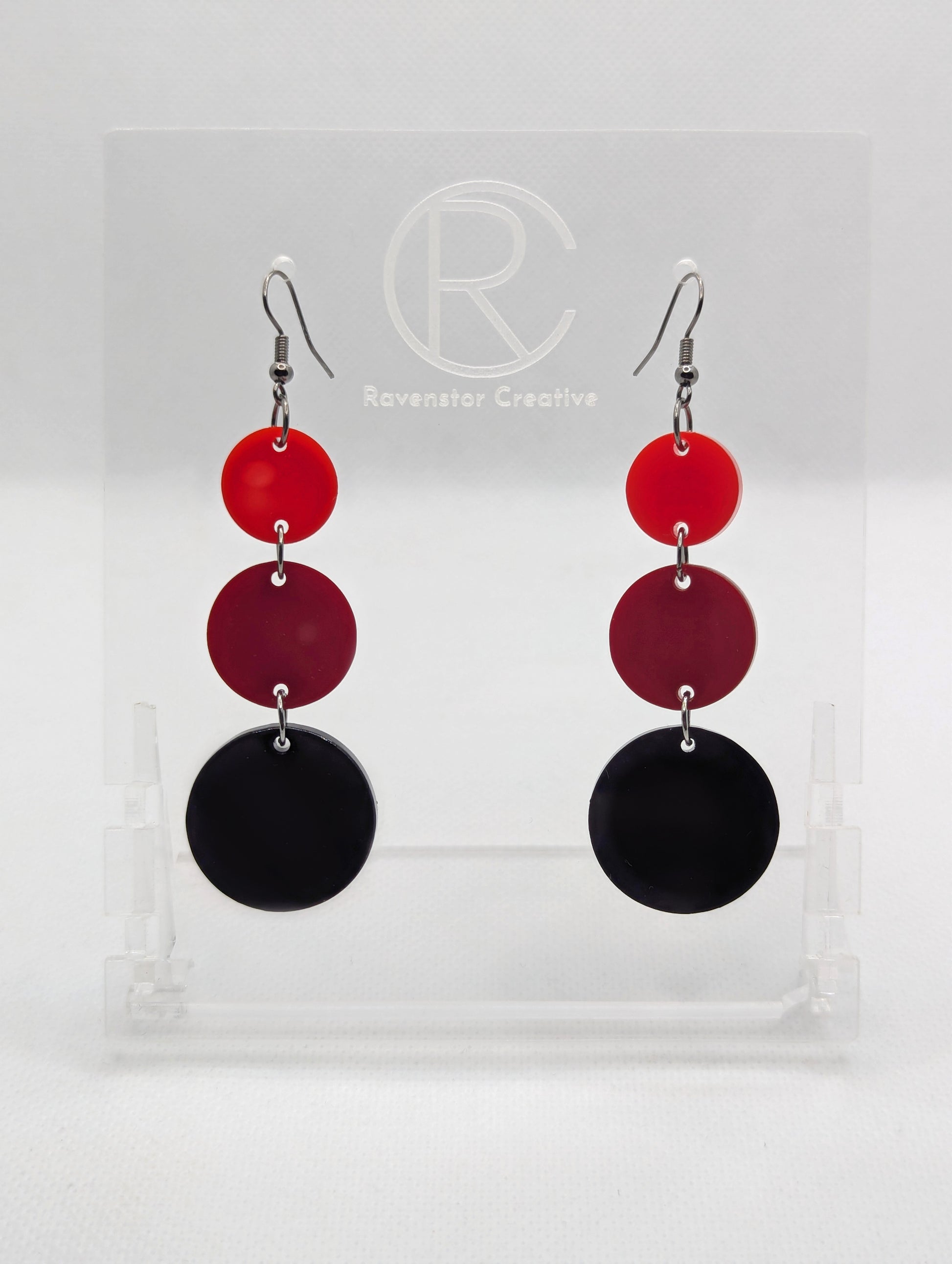 A pair of acrylic earrings with black ear wire hook fixings. Each has three circles dangling from each other, the top is the smallest and they get bigger as they go down. The colours from the top are Red, Maroon and Black.