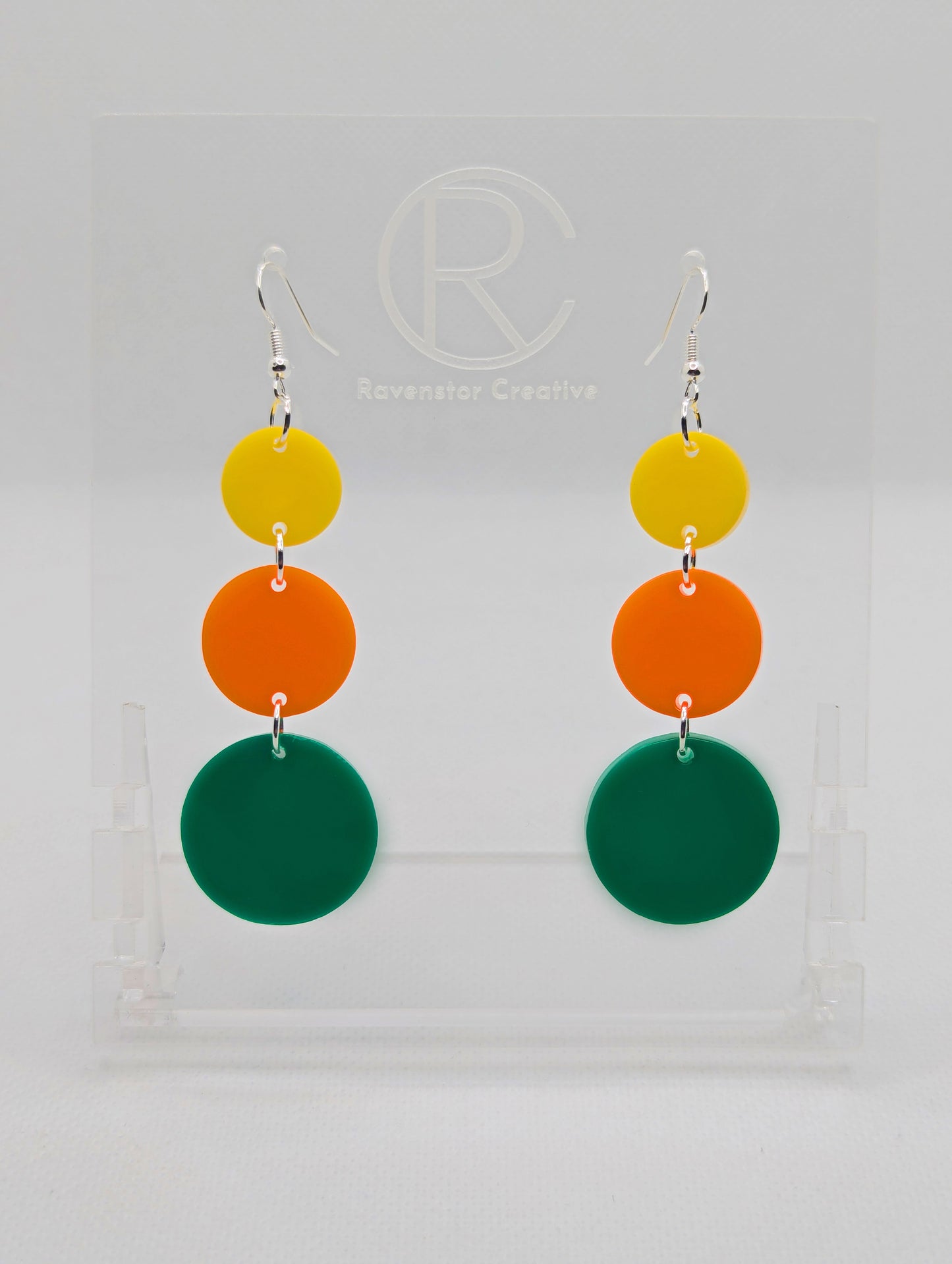 A pair of acrylic earrings with Silver ear wire hook fixings. Each has three circles dangling from each other, the top is the smallest and they get bigger as they go down. The colours from the top are Yellow, Orange and Green.