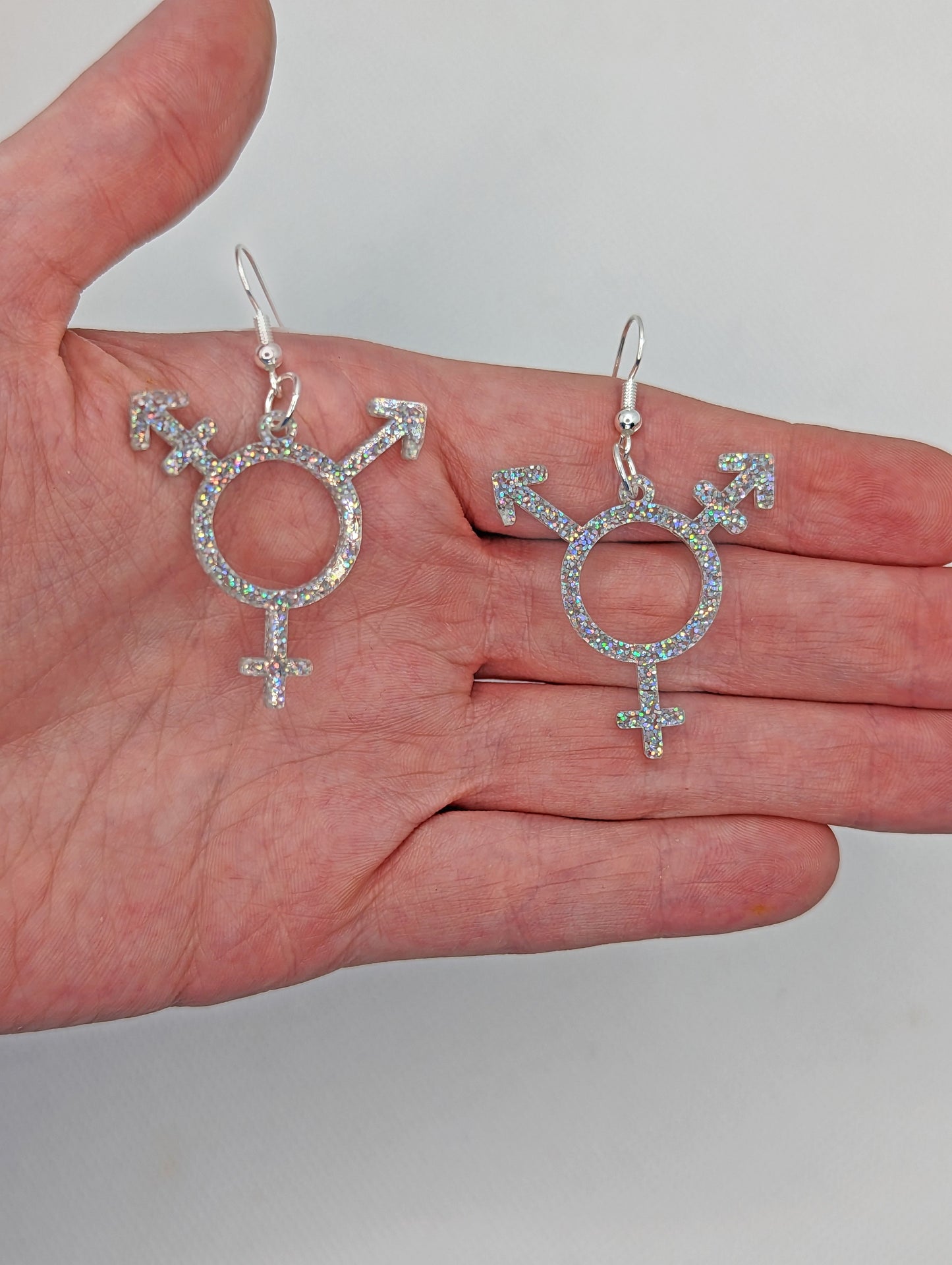 A pair of acrylic earrings in the shape of the Pride Transgender LGBTQ symbol. They have ear wire hook fixings and are in Holographic Glitter.  They are against a hand to show the size of 2 and a half finger widths length and the same again wide.