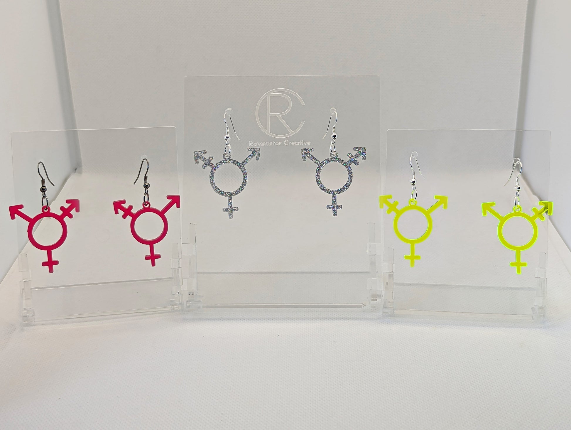 Three pairs of acrylic earrings in the shape of the Pride Transgender   LGBTQ symbol. They have ear wire hook fixings and the colours are Hot Pink, Holographic Glitter and Neon Green. They are on clear stands against a white background.