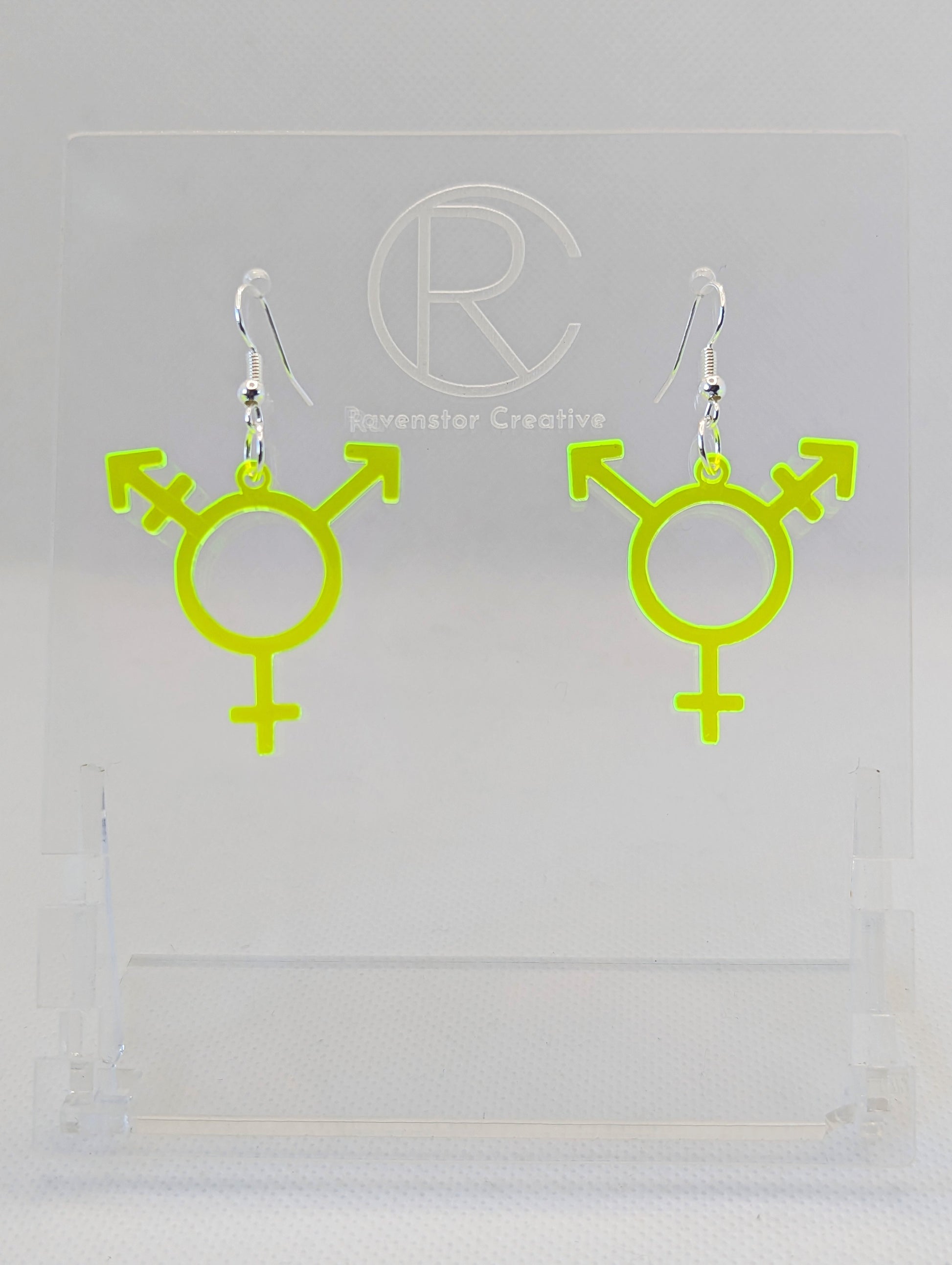 A pair of acrylic earrings in the shape of the Pride Transgender LGBTQ symbol. They have Silver ear wire hook fixings and the colour is Neon Green.  They are on a clear stand with the Ravenstor Creative Logo on it against a white background.