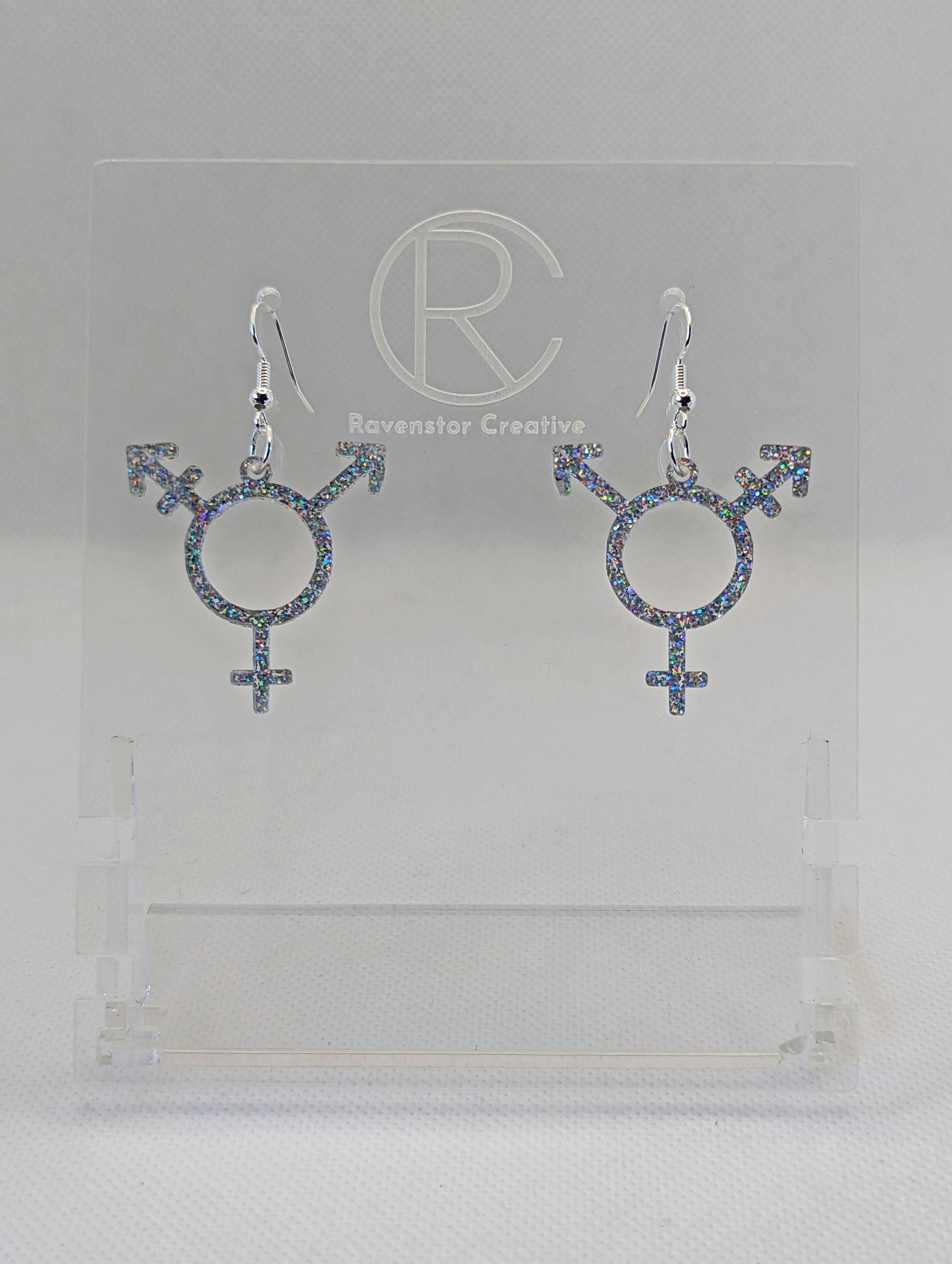 A pair of acrylic earrings in the shape of the Pride Transgender LGBTQ symbol. They have Silver ear wire hook fixings and the colour is Holographic Glitter.  They are on a clear stand with the Ravenstor Creative Logo on it against a white background.