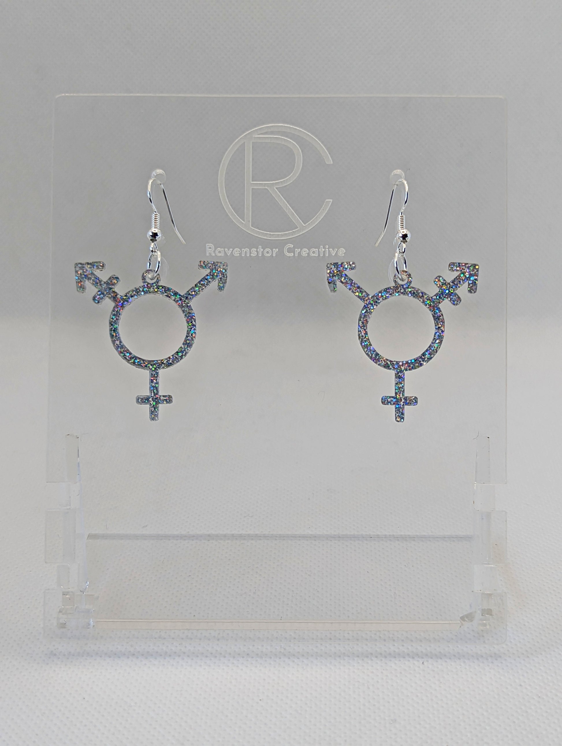 A pair of acrylic earrings in the shape of the Pride Transgender LGBTQ symbol. They have Silver ear wire hook fixings and the colour is Holographic Glitter.  They are on a clear stand with the Ravenstor Creative Logo on it against a white background.