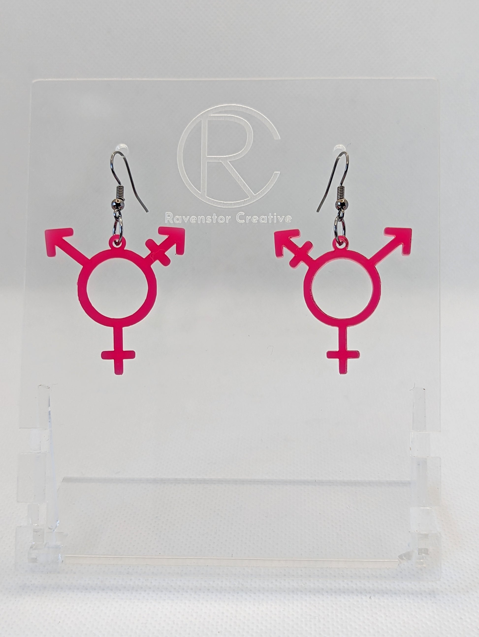 A pair of acrylic earrings in the shape of the Pride Transgender LGBTQ symbol. They have Black ear wire hook fixings and the colour is Hot Pink.  They are on a clear stand with the Ravenstor Creative Logo on it against a white background.