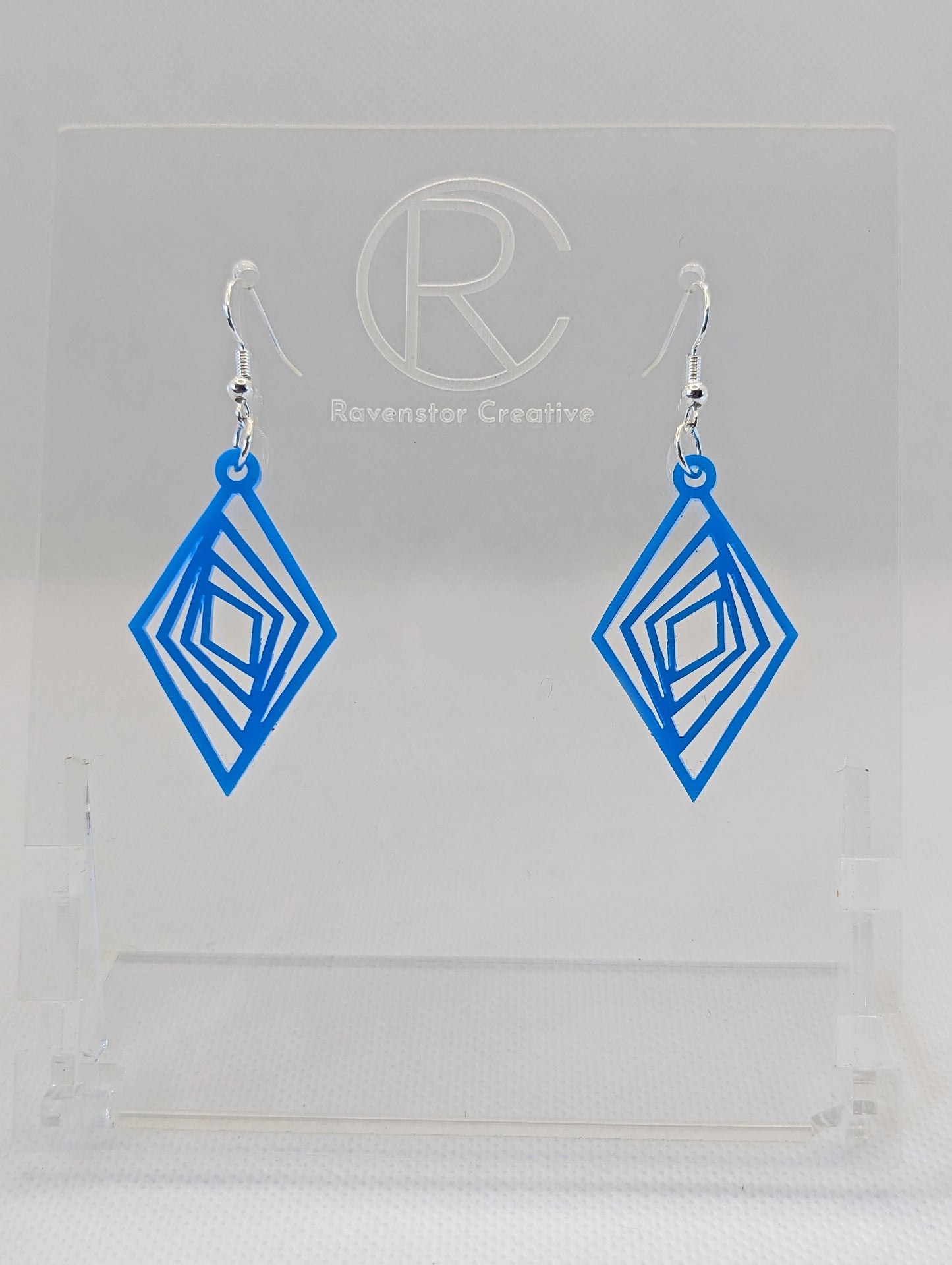 These are acrylic diamond shape earrings with twisted Geometric patterns cut into the middle. They have silver ear wire hook fixings and they are Blue in colour. They are on a clear stand against a white background.