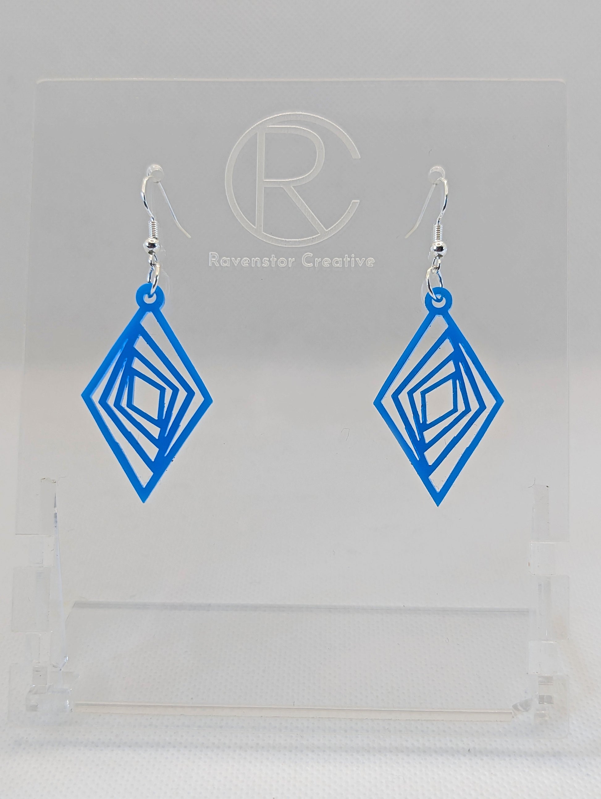 These are acrylic diamond shape earrings with twisted Geometric patterns cut into the middle. They have silver ear wire hook fixings and they are Blue in colour. They are on a clear stand against a white background.