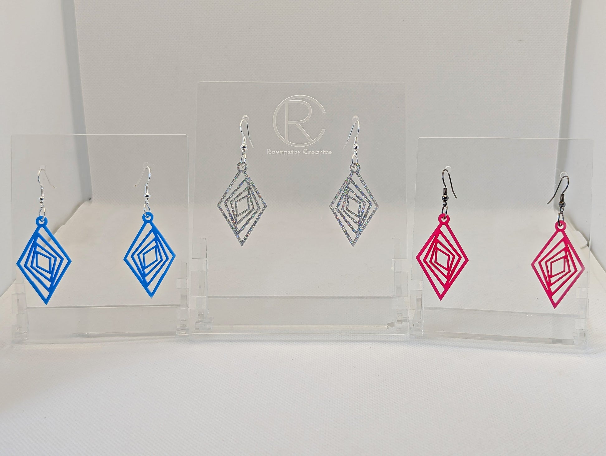 Three pairs of acrylic diamond shape earrings with twisted Geometric patterns cut into the middle. They have ear wire hook fixings and the colours are Blue, Holographic Glitter and Hot Pink. They are on clear stands against a white background.