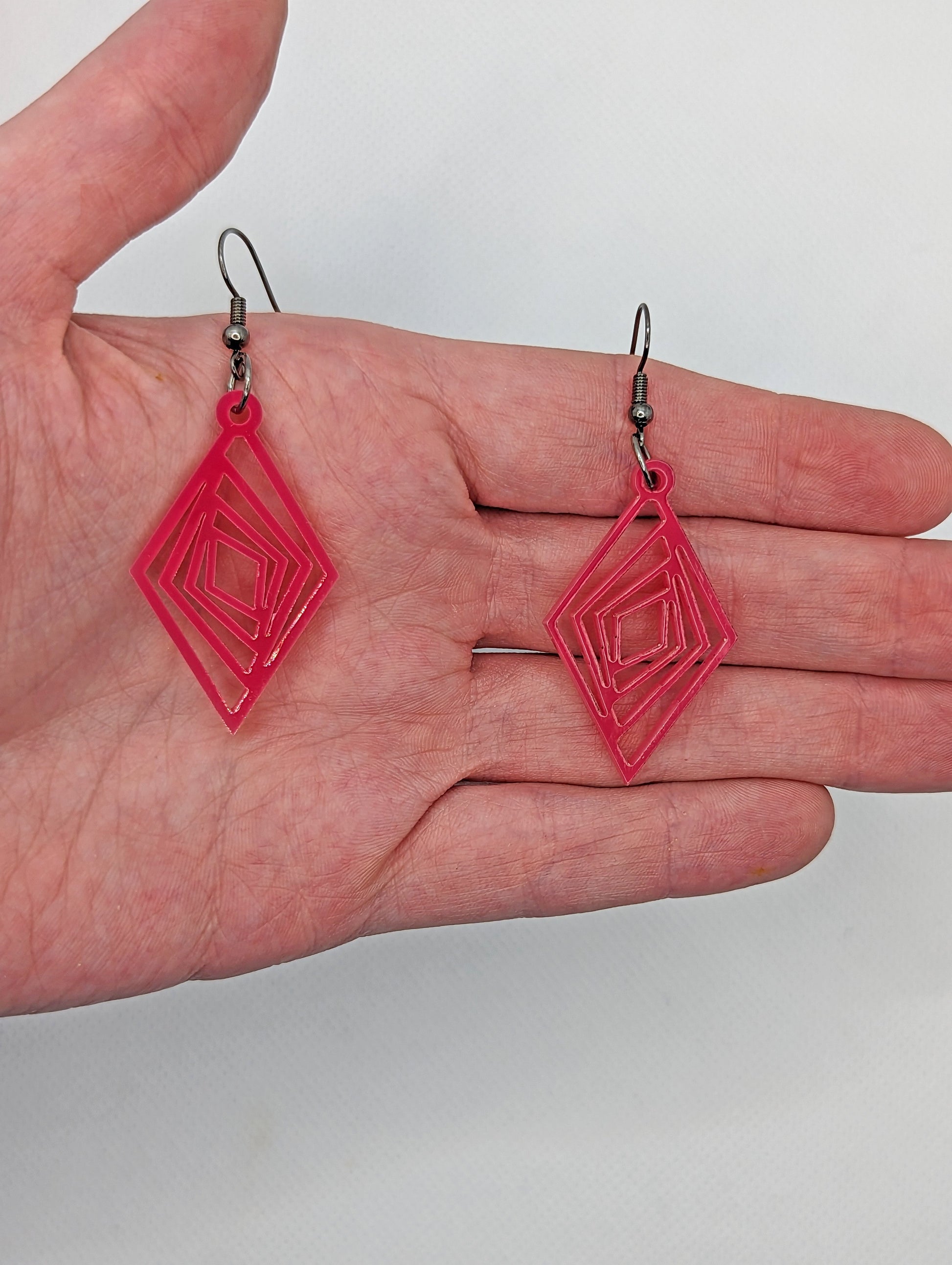 A pair of acrylic diamond shape earrings with twisted Geometric patterns cut into the middle. They have ear wire hook fixings and are against a hand to show the size.  They are two and a half finger widths in length and one and a half wide.