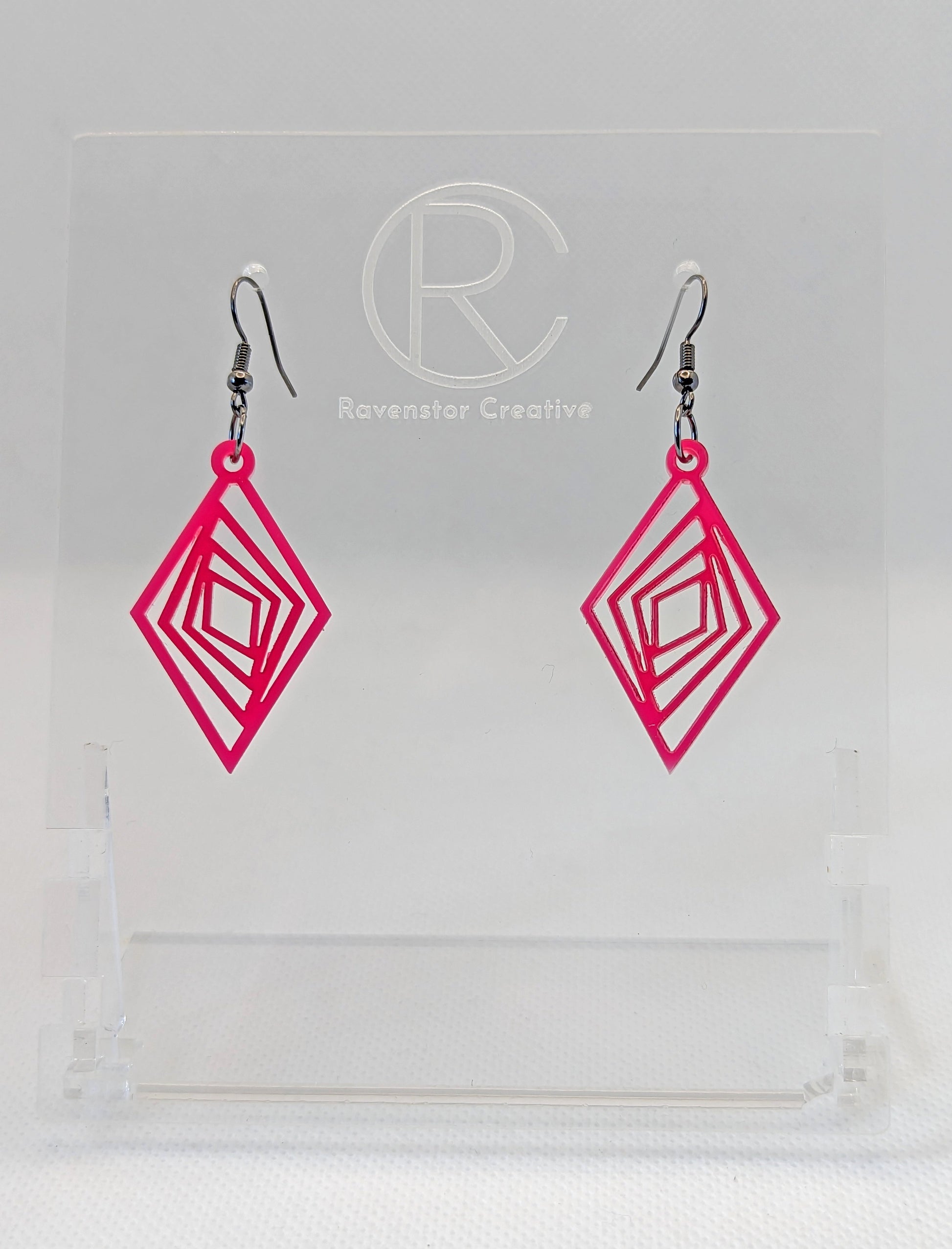 These are acrylic diamond shape earrings with twisted Geometric patterns cut into the middle. They have black ear wire hook fixings and they are hot pink in colour. They are on a clear stand against a white background.