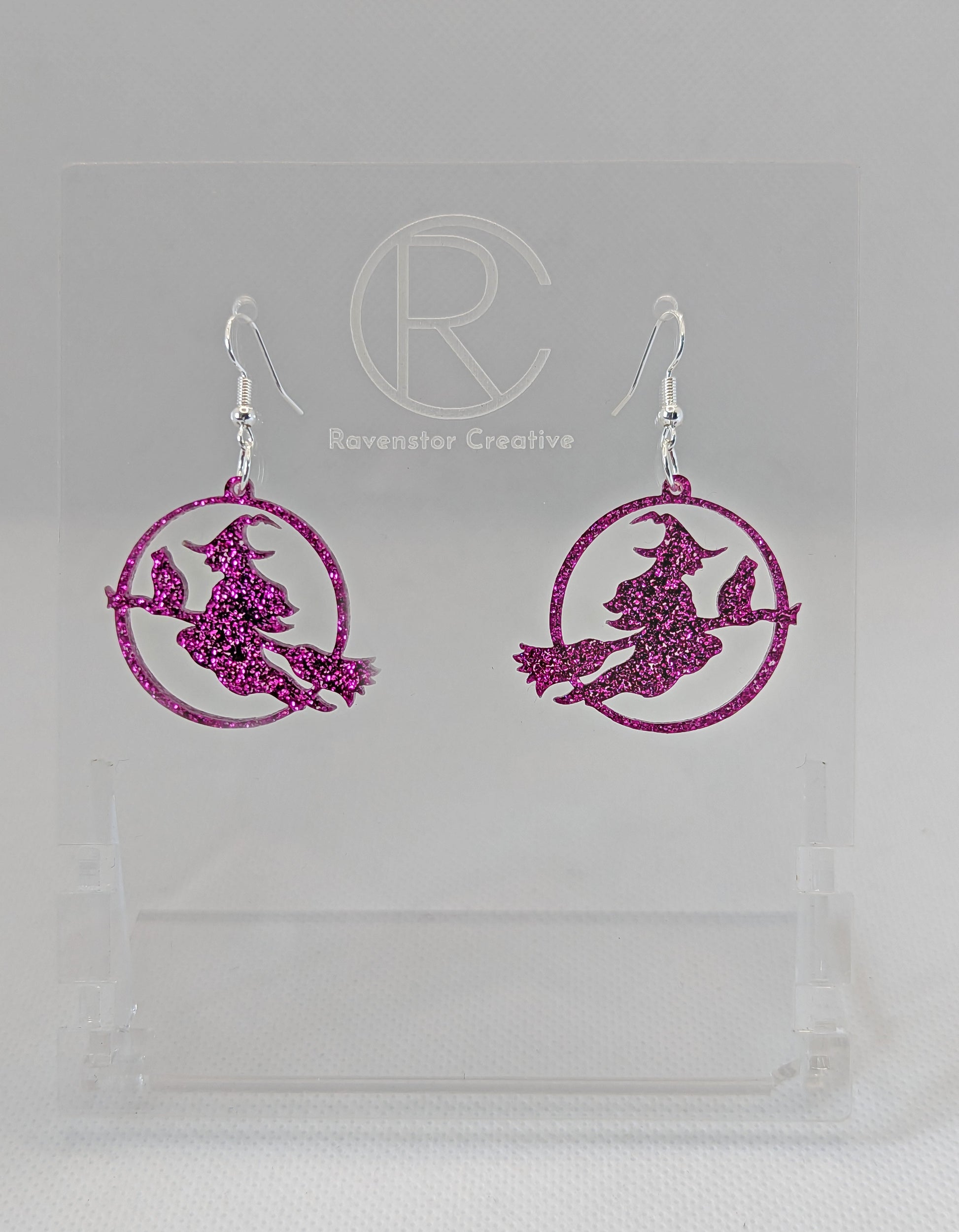 A pair of acrylic earrings with silver Ear wire hook fixings on a white background, they are a circle with a scene cut out of a witch on her broom with a cat perched on the broom. The colour is a pretty pink glitter.