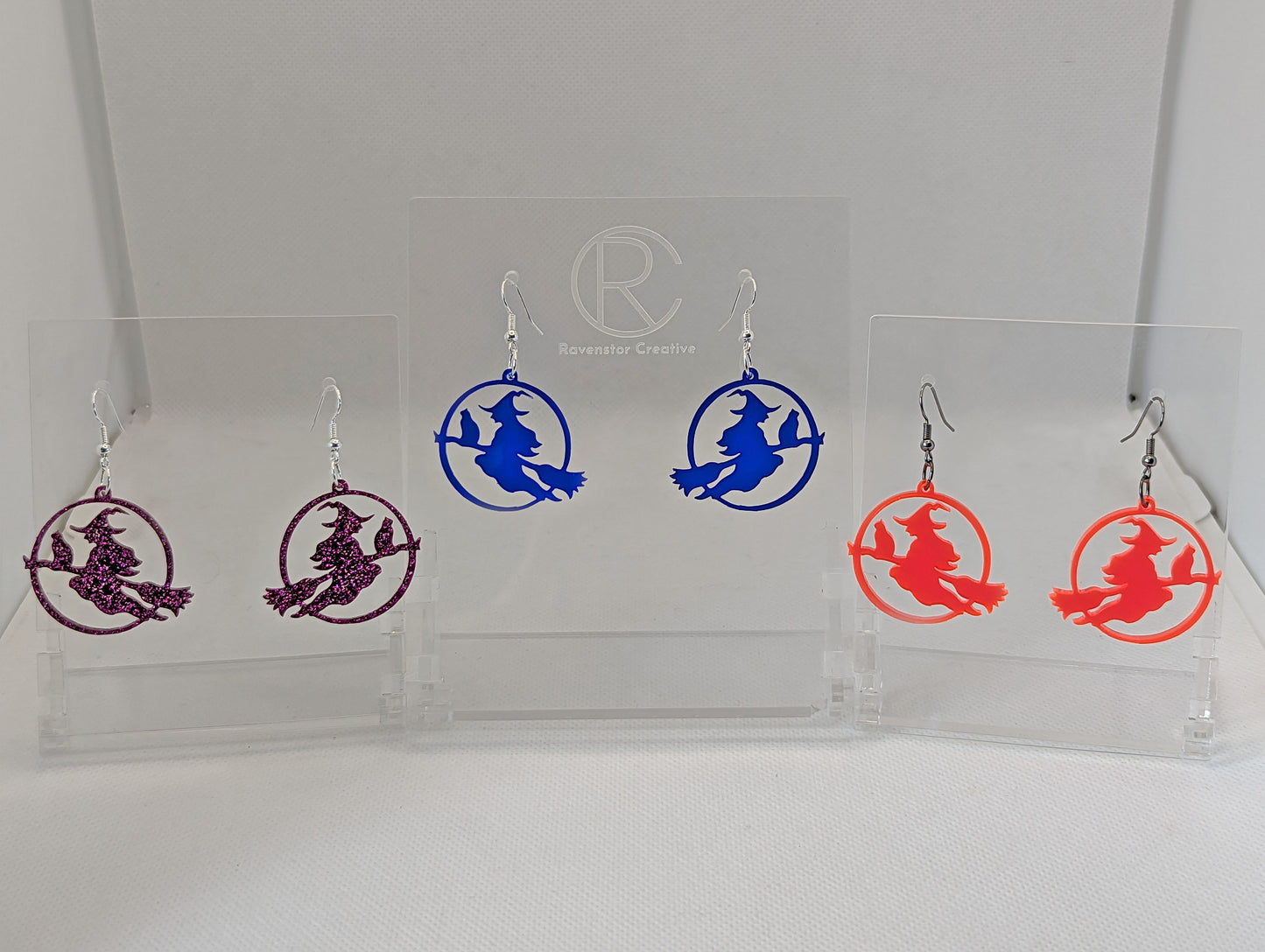 An image of 3 pairs of earrings with silver & black fixings on a white background, they are a circle with a scene cut out of a witch on her broom with a cat perched on the broom. The colours shown are glittery pink, translucent blue and neon pink.