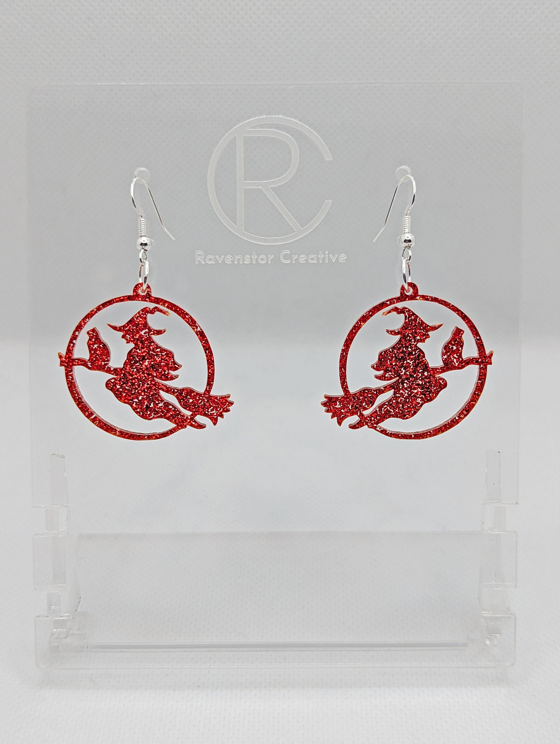 A pair of acrylic earrings with silver Ear wire hook fixings on a white background, they are a circle with a scene cut out of a witch on her broom with a cat perched on the broom. The colour is a lovely Red Glitter.