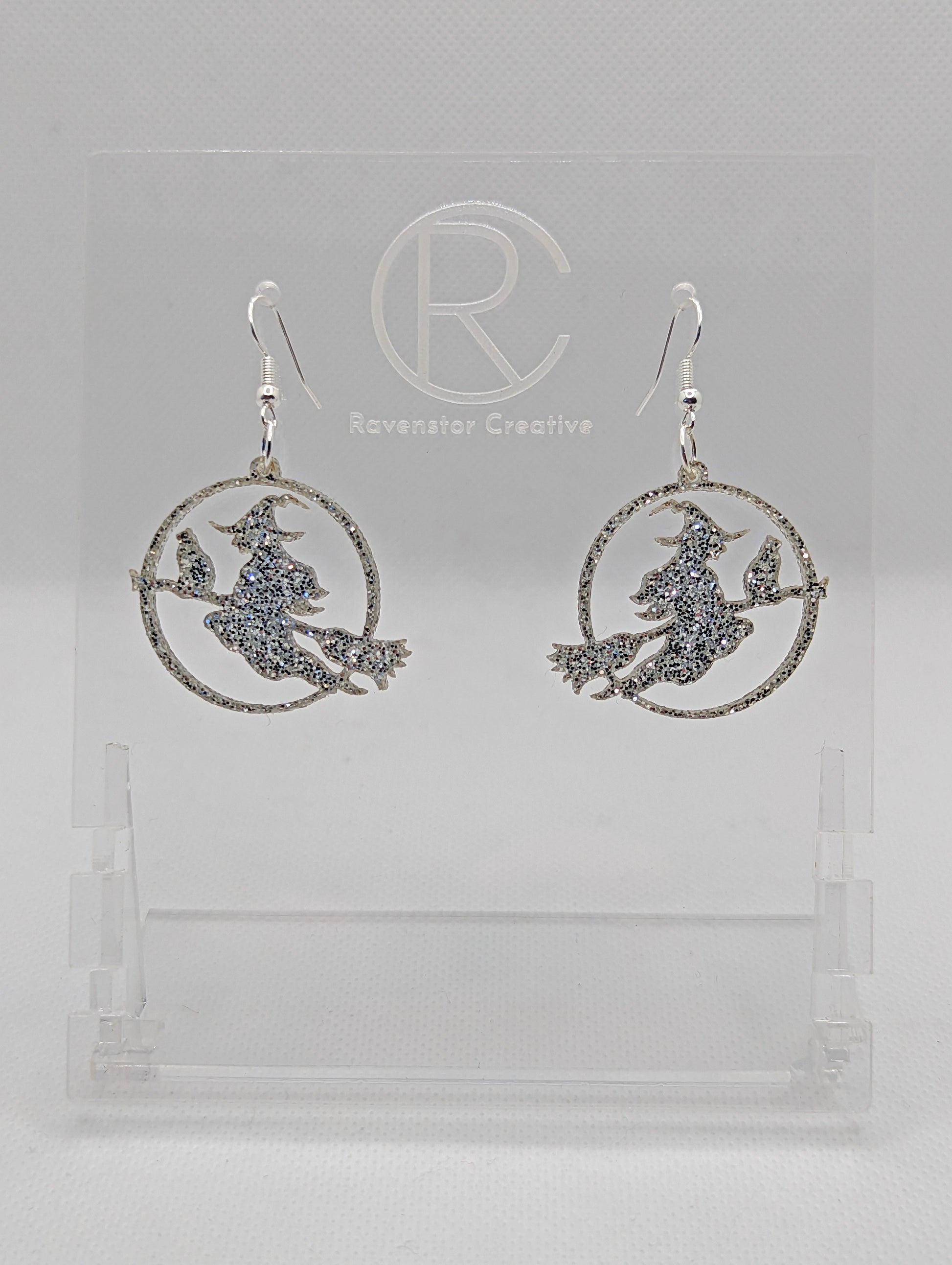 A pair of acrylic earrings with silver Ear wire hook fixings on a white background, they are a circle with a scene cut out of a witch on her broom with a cat perched on the broom. The colour is a pretty Silver Glitter.