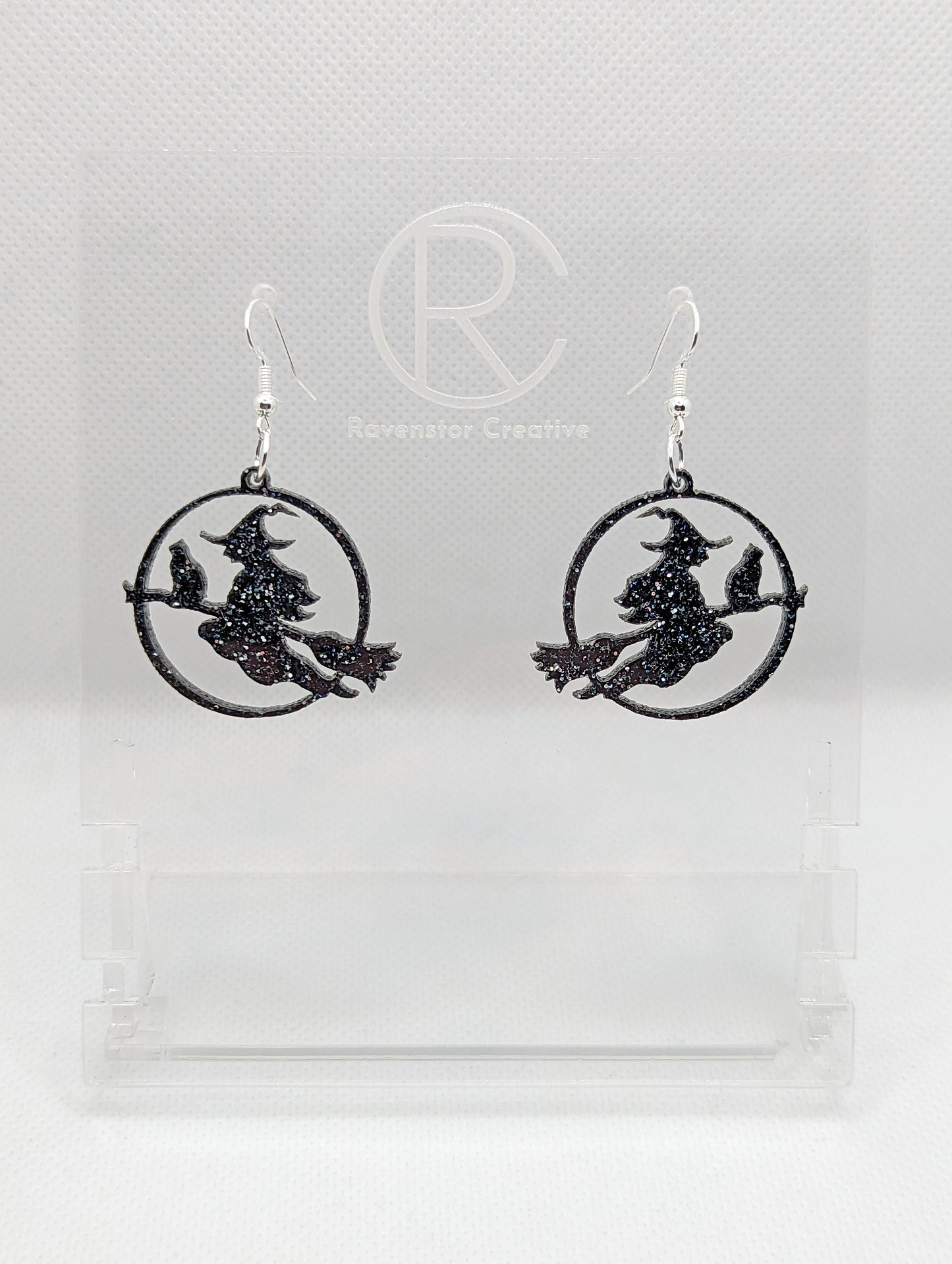 A pair of acrylic earrings with silver Ear wire hook fixings on a white background, they are a circle with a scene cut out of a witch on her broom with a cat perched on the broom. The colour is a lovely Black and Silver Glitter.