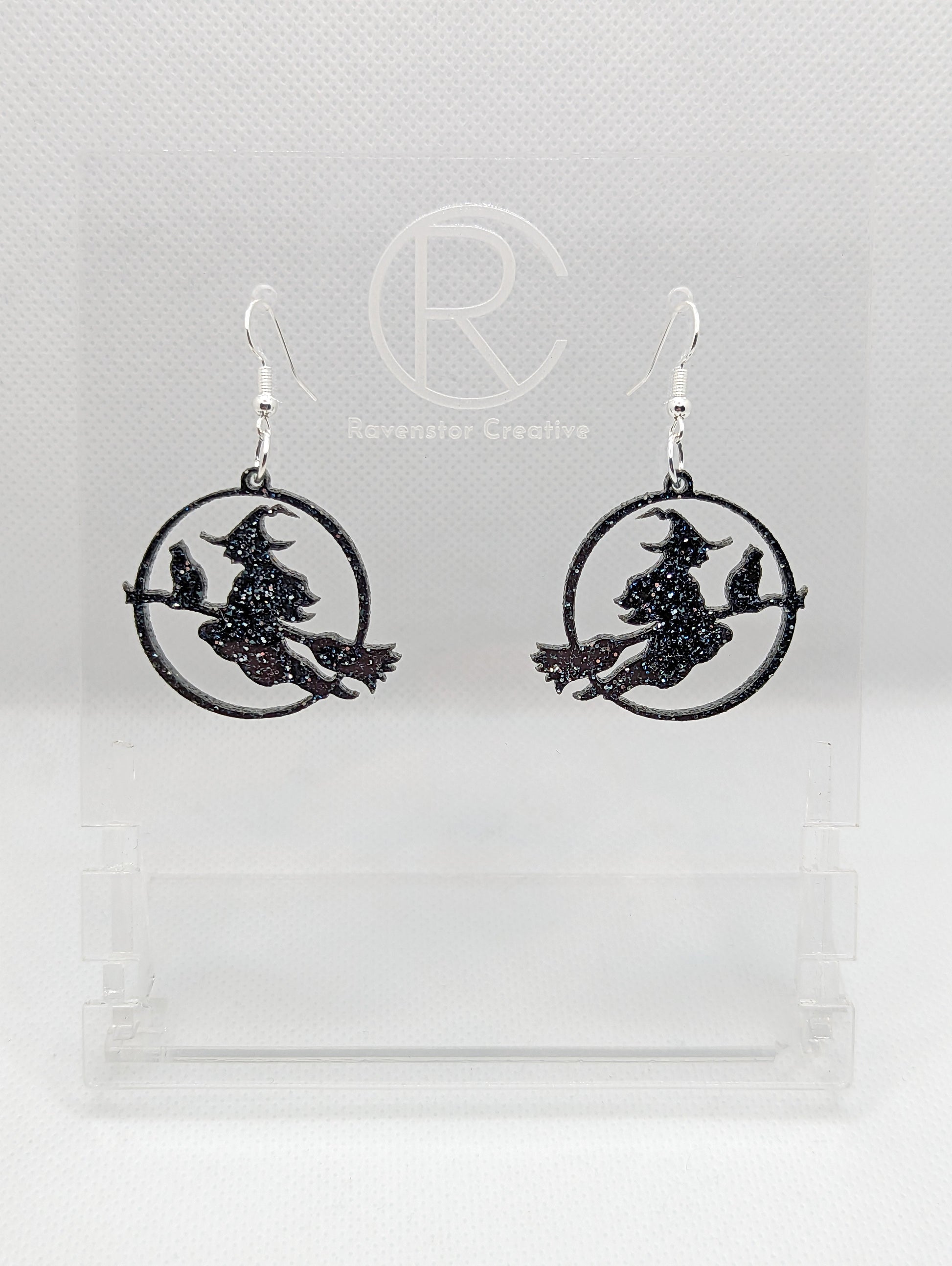 A pair of acrylic earrings with silver Ear wire hook fixings on a white background, they are a circle with a scene cut out of a witch on her broom with a cat perched on the broom. The colour is a lovely Black and Silver Glitter.