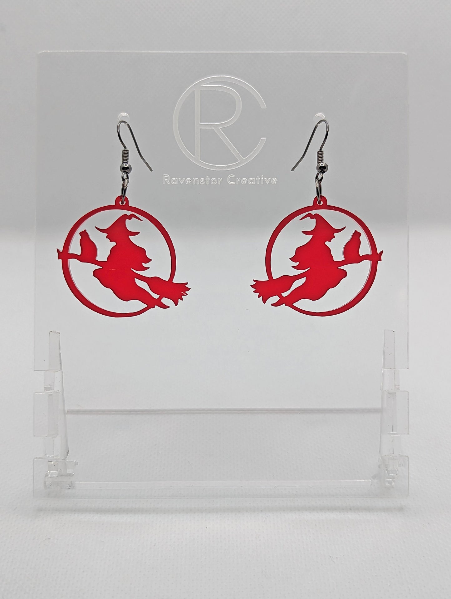 A pair of acrylic earrings with black Ear wire hook fixings on a white background, they are a circle with a scene cut out of a witch on her broom with a cat perched on the broom. The colour is a spooky translucent Red.