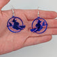 A pair of earrings in a hand to show the size with silver fixings, they are a circle with a scene cut out of a witch on her broom with a cat perched on the broom. They are translucent blue, the earrings themselves are about 2 fingers width and length