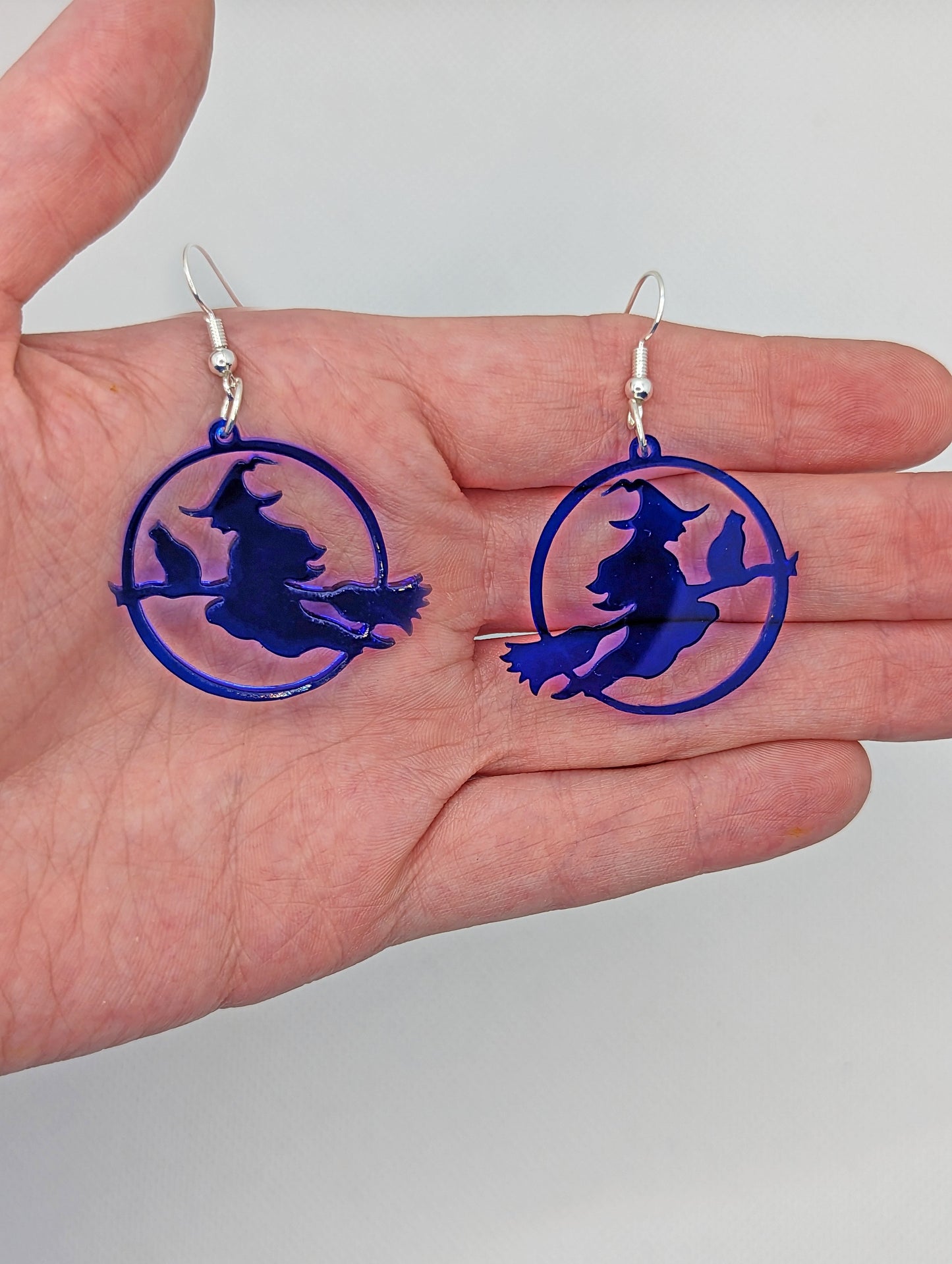 A pair of earrings in a hand to show the size with silver fixings, they are a circle with a scene cut out of a witch on her broom with a cat perched on the broom. They are translucent blue, the earrings themselves are about 2 fingers width and length