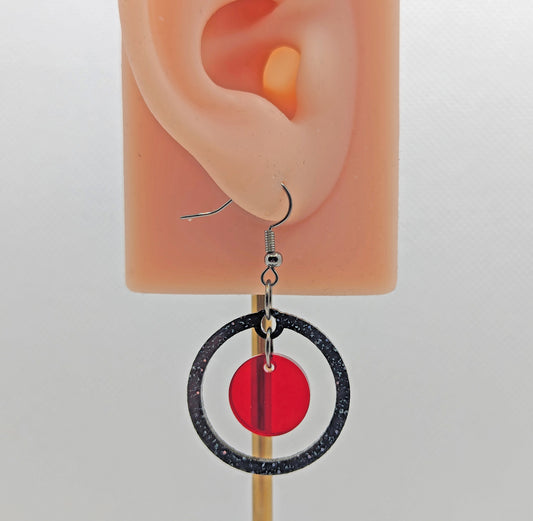 A pair of circle shaped earrings with a smaller circle dangling inside on an ear showing thier size. They are Black & Silver Glitter for the outer circle and Translucent Red for the small inner Circle.