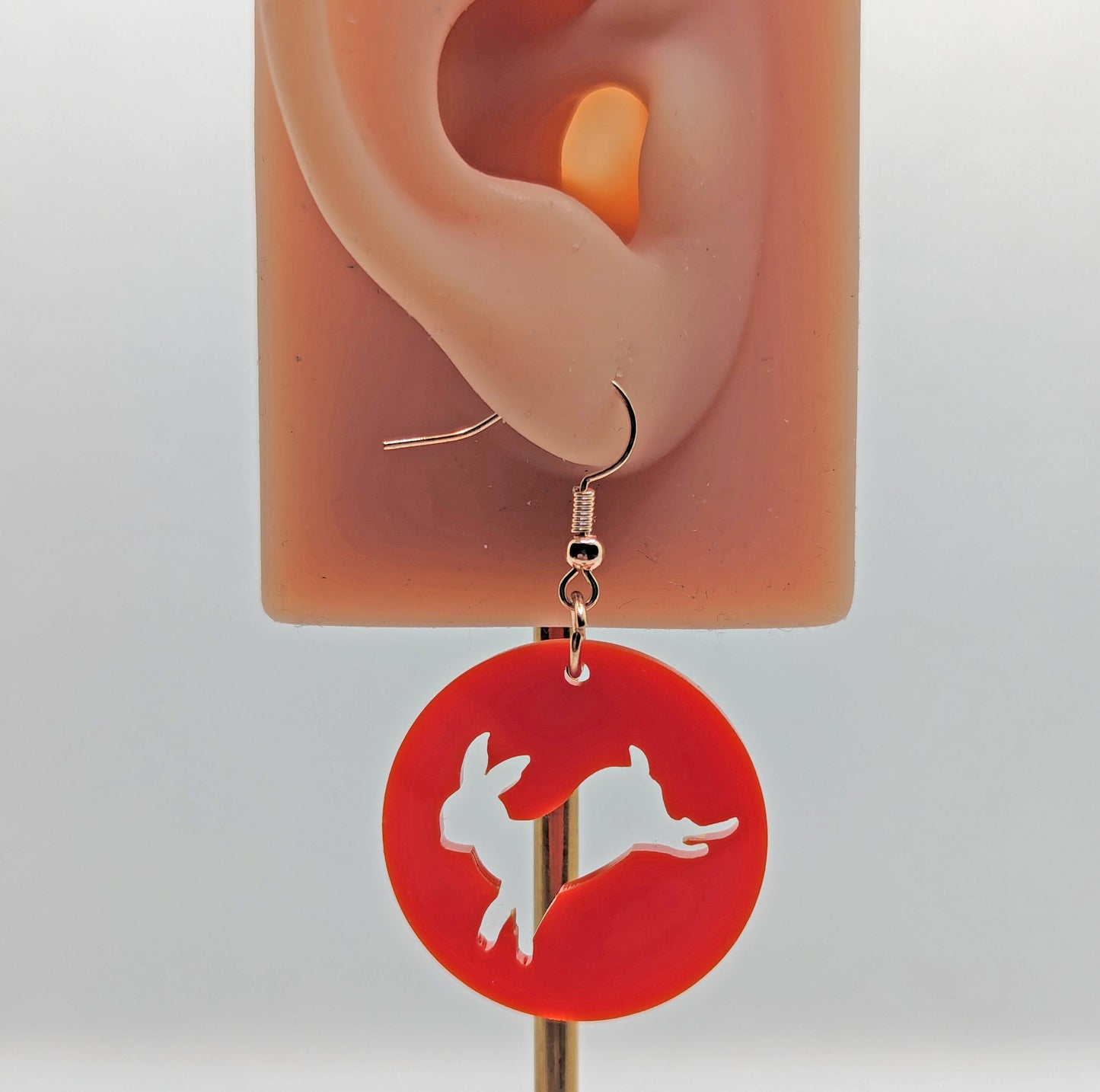 An acrylic earring that has rose gold ear wire hook fixings against an ear to show the size. It is a Circle with an Easter bunny / Rabbit hopping cut out of it. It is Red in colour.