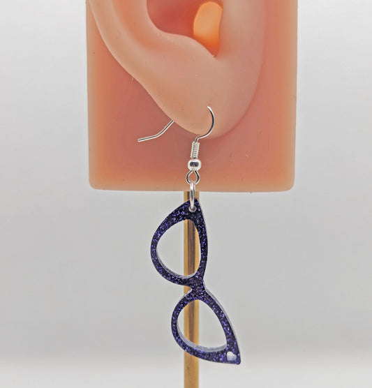 Image of an of fab sassy glasses earrings on an ear showing the size against a white background. It is purple glitter in colour and have silver fixings.