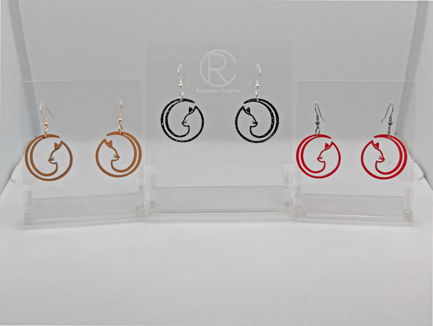 Shows three pairs of earrings that have ear wire hook fixings. They are full moon cat imspired. The colours are Cherry Wood, Black & Silver Glitter and Translucent Red. They are against a white background on see through stands.