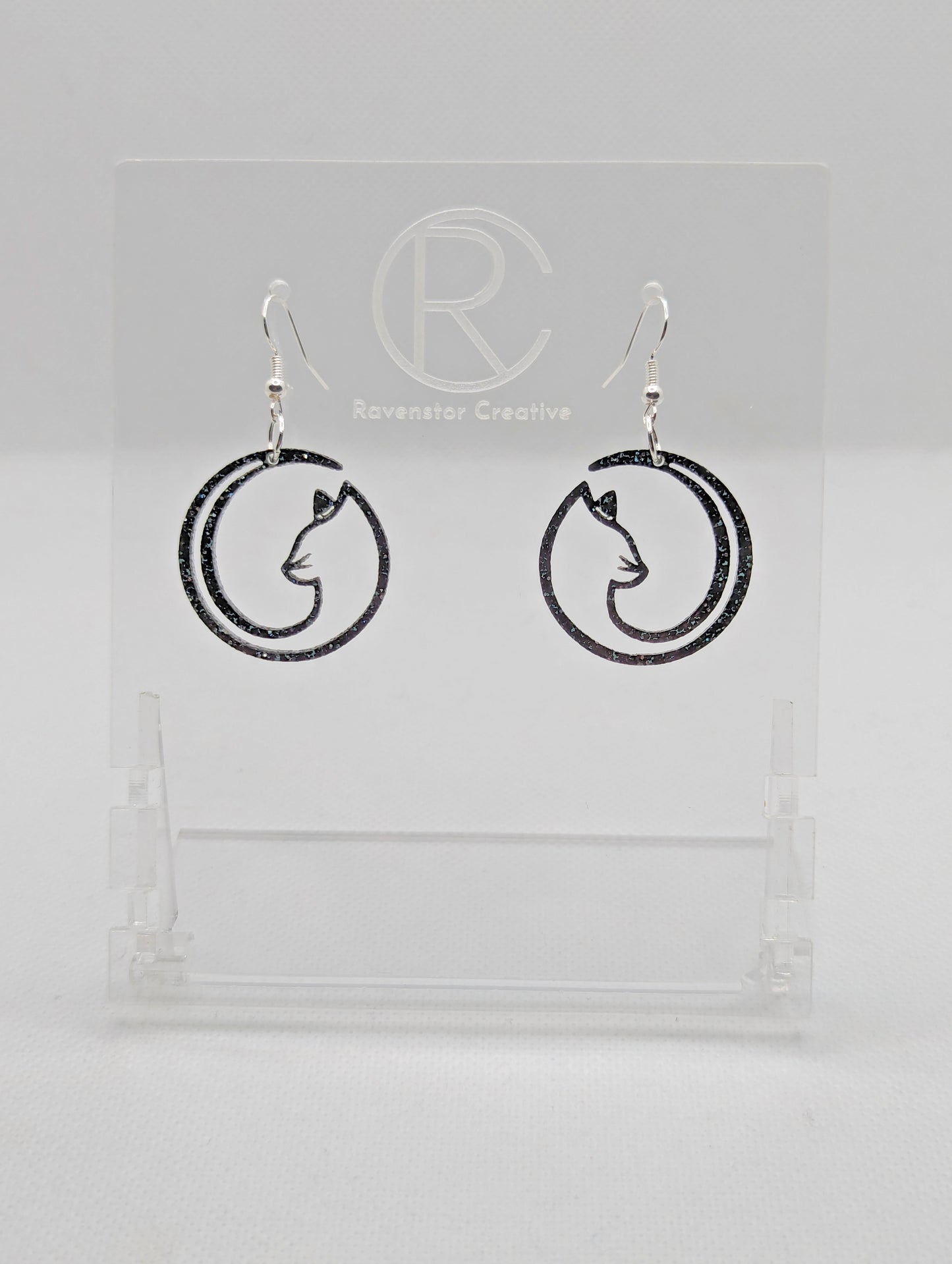 Shows a pair of acrylic earrings that have silver ear wire hook fixings. They are Full Moon inspired Cat shapes. They are in Purple Glitter and are against a white background on see through stand with the Ravenstor Creative logo on it.