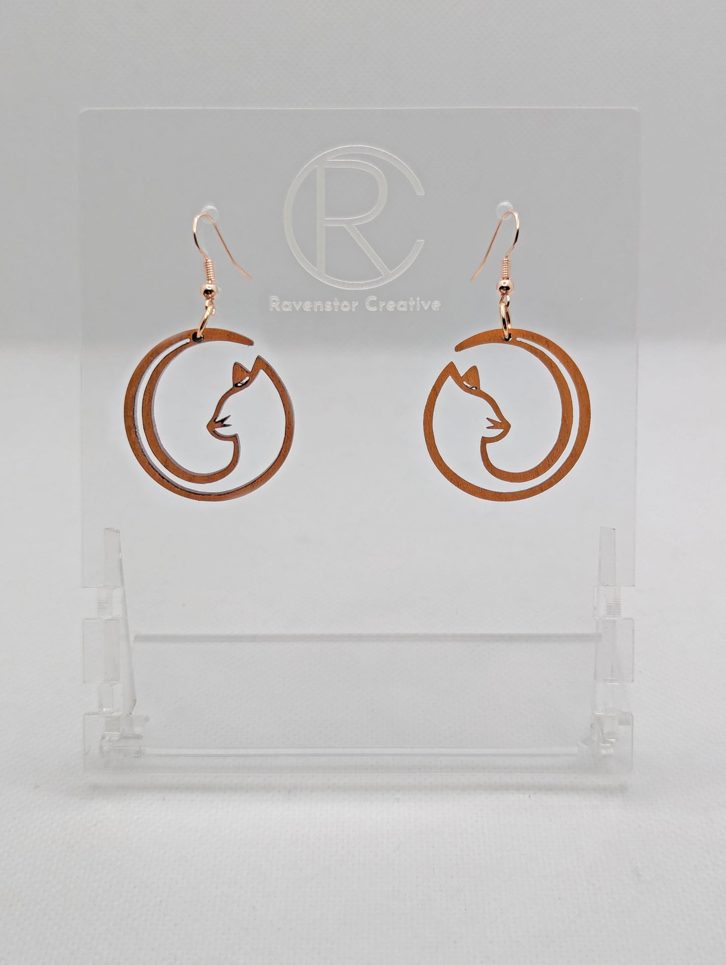 Shows a pair of earrings that have rose gold ear wire hook fixings. They are Full Moon inspired Cat shapes. They are in Cherry Wood and are against a white background on see through stand with the Ravenstor Creative logo on it.