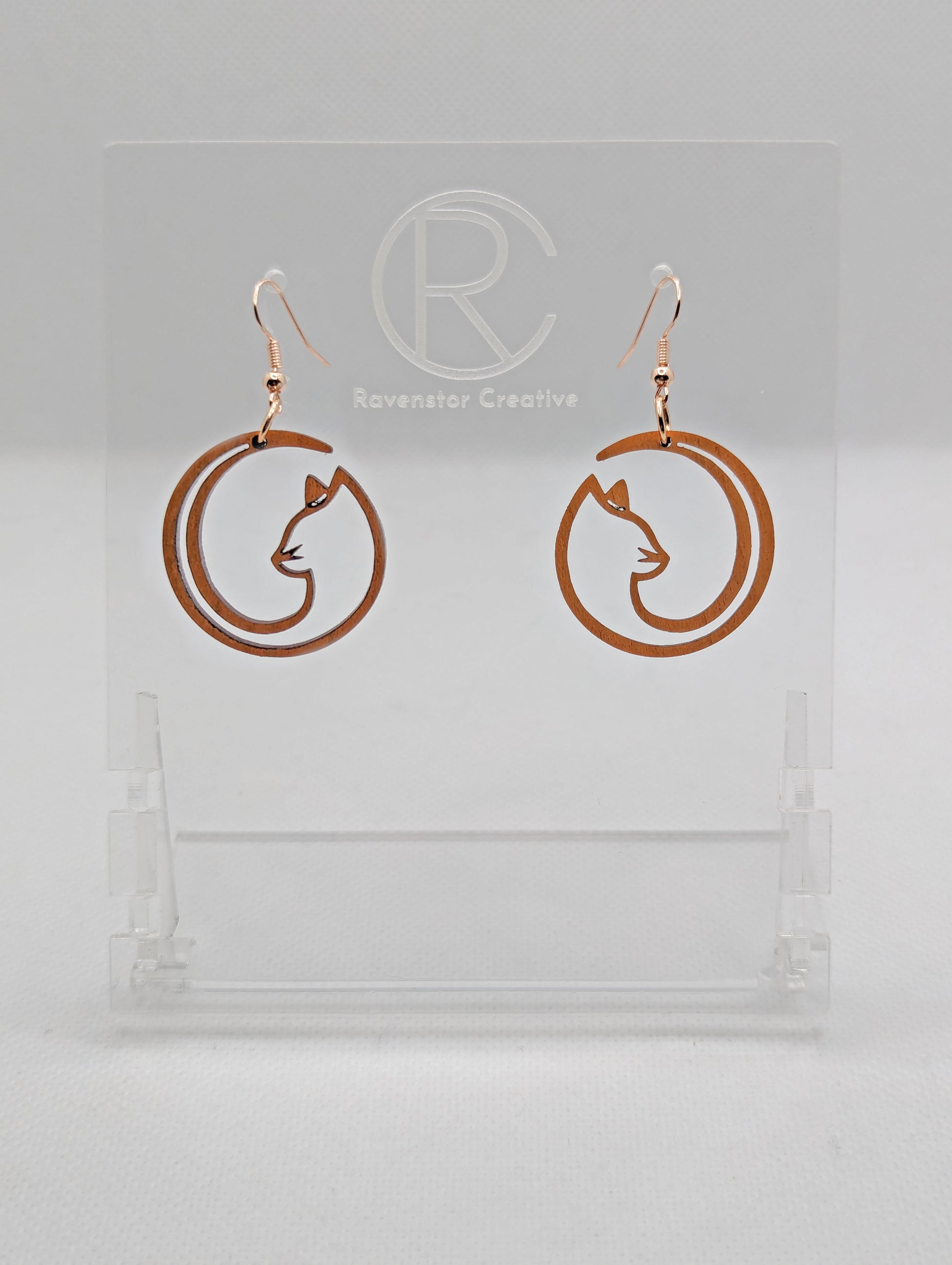 Shows a pair of earrings that have rose gold ear wire hook fixings. They are Full Moon inspired Cat shapes. They are in Cherry Wood and are against a white background on see through stand with the Ravenstor Creative logo on it.