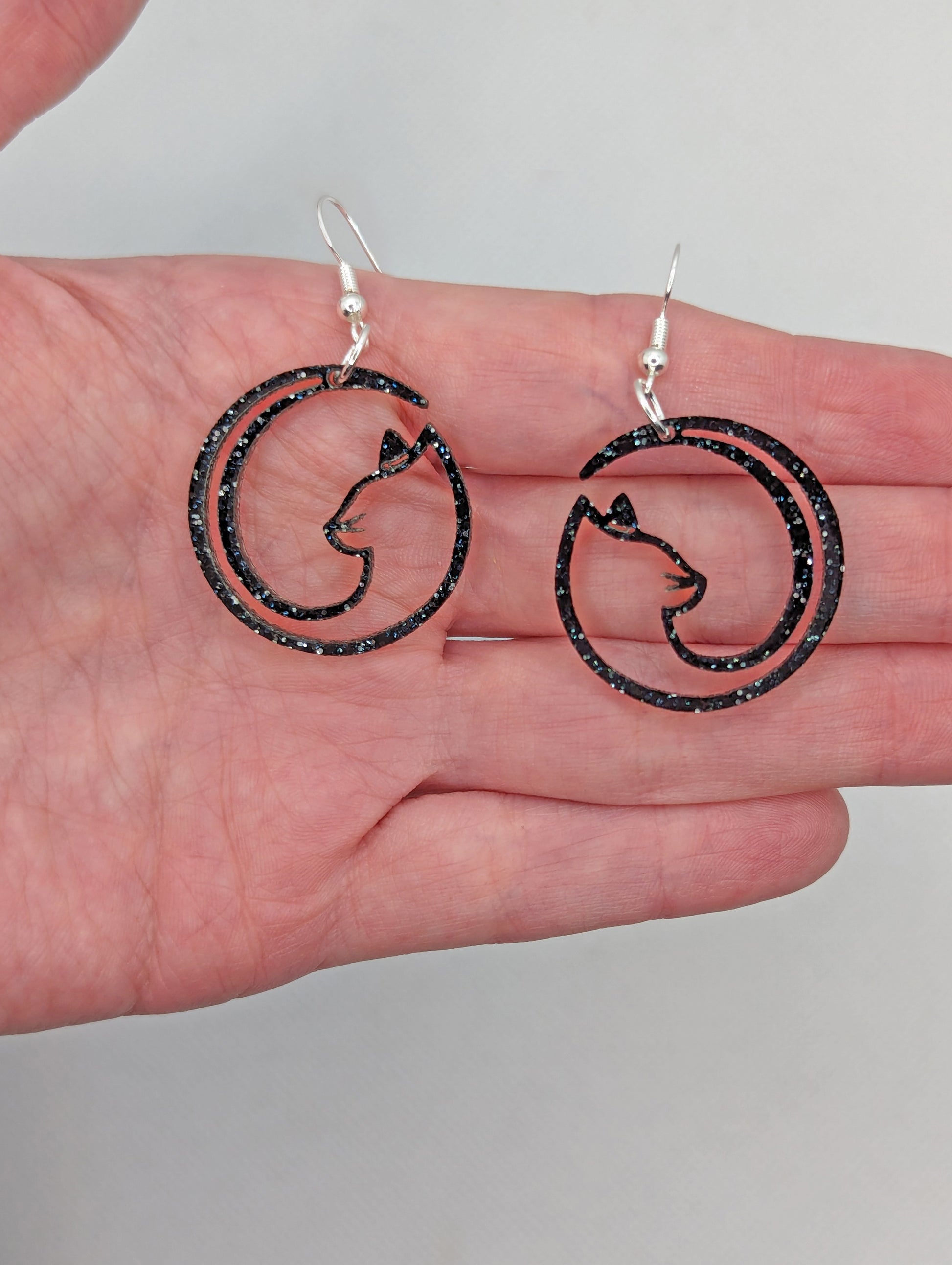 Image of a pair of earrings that are full moon inspired cat shapes, on a hand showing the size against a white background, they are 2 finger widths length the same again wide. They are black and silver glitter and have silver fixings.