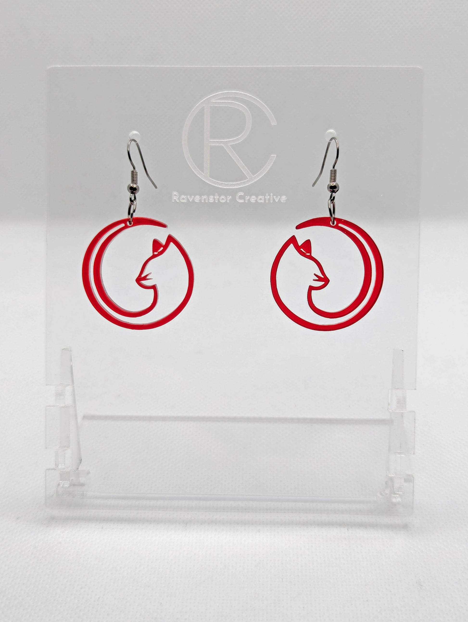 Shows a pair of acrylic earrings that have black ear wire hook fixings. They are Full Moon inspired Cat shapes. They are in translucent red and are against a white background on see through stand with the Ravenstor Creative logo on it.