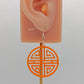 Shows a Geometric Korean Inspired Circle earring with an intricate pattern like a maze. Its on an ear showing the size against a white background. They are orange acrylic and have rose gold fixings.