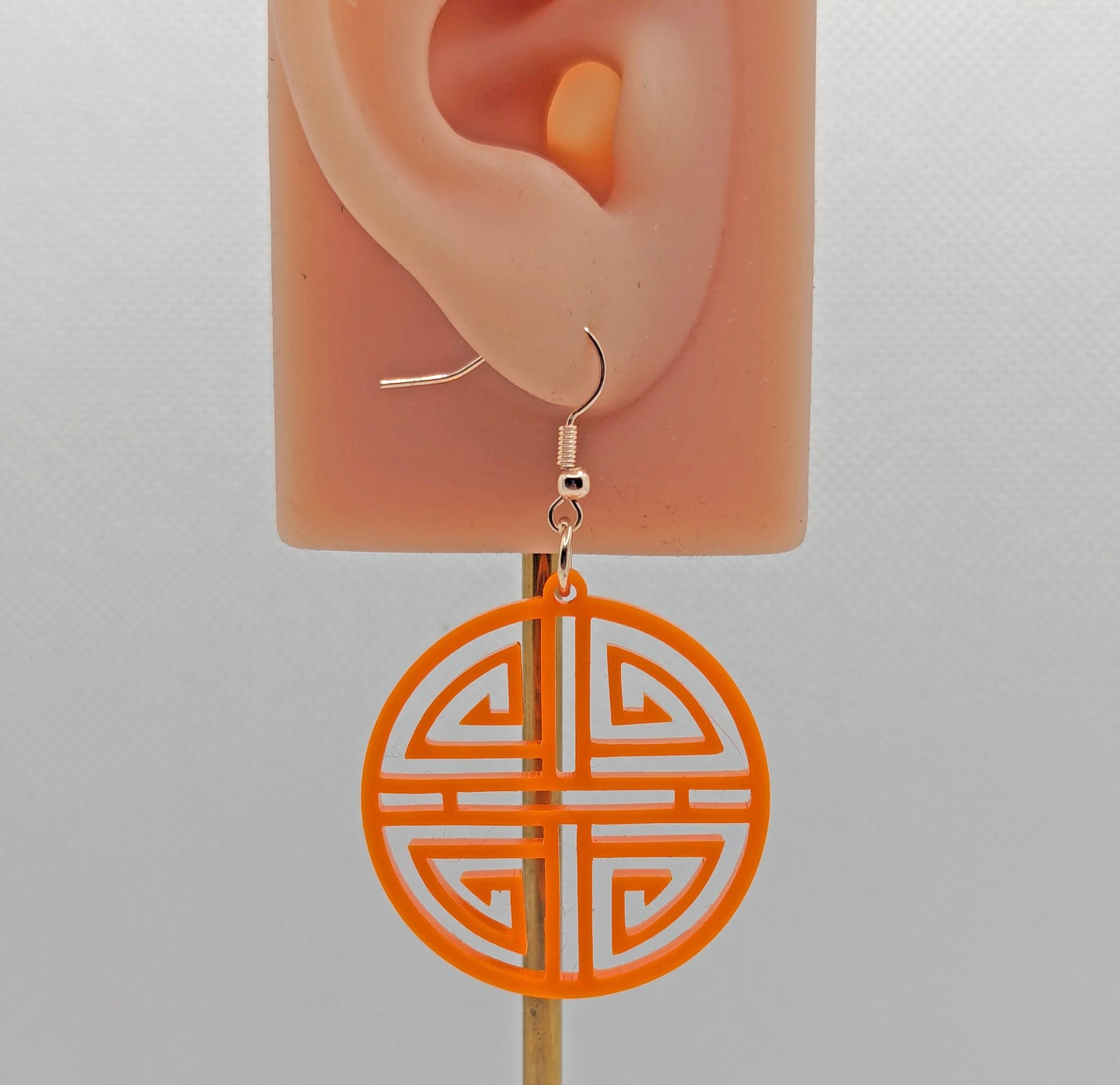 Shows a Geometric Korean Inspired Circle earring with an intricate pattern like a maze. Its on an ear showing the size against a white background. They are orange acrylic and have rose gold fixings.