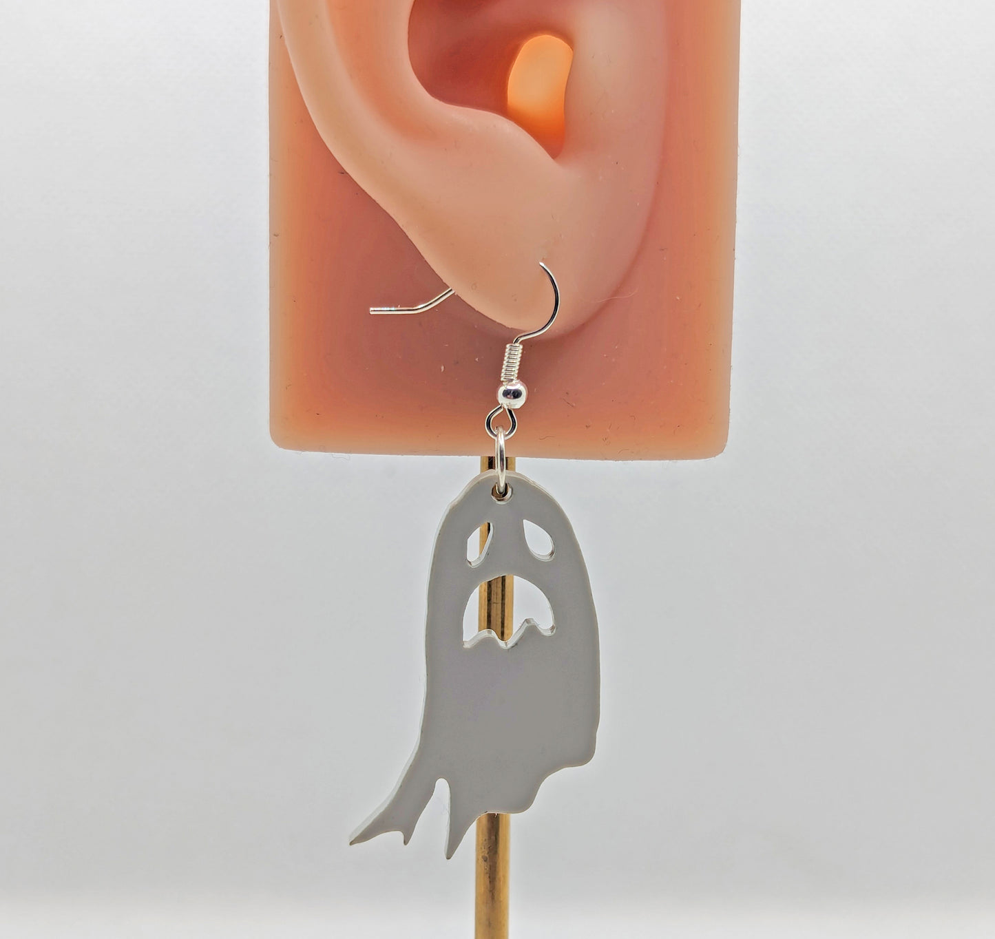 Image of a spooky moaning Ghost shaped earring on an ear showing the size against a white background. They are grey and have silver ear wire hook fixings.