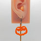 Image of a scary Halloween pumpkin shaped earring on an ear showing the size against a white background. They are orange acrylic and have black fixings.