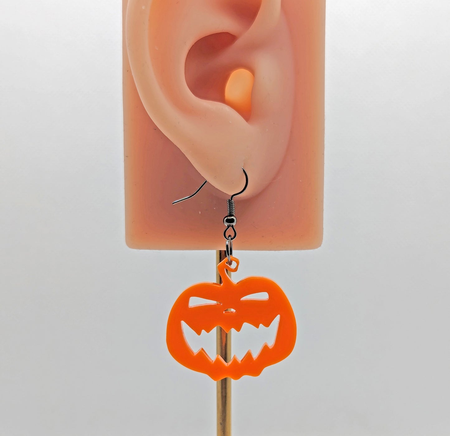 Image of a scary Halloween pumpkin shaped earring on an ear showing the size against a white background. They are orange acrylic and have black fixings.