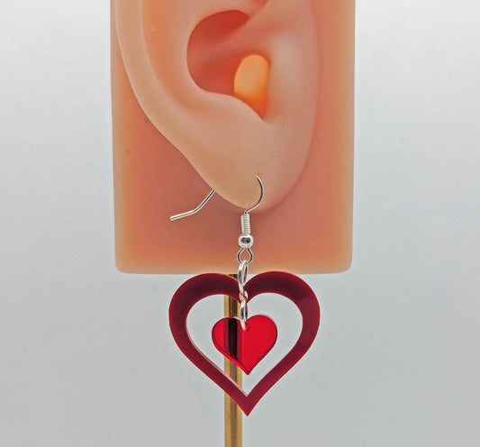 Image of a heart shaped earring with a smaller heart dangling inside on an ear showing thier size. It is maroon acrylic  for the outer bigger heart and translucent red for the small inner heart. It is against a white background.