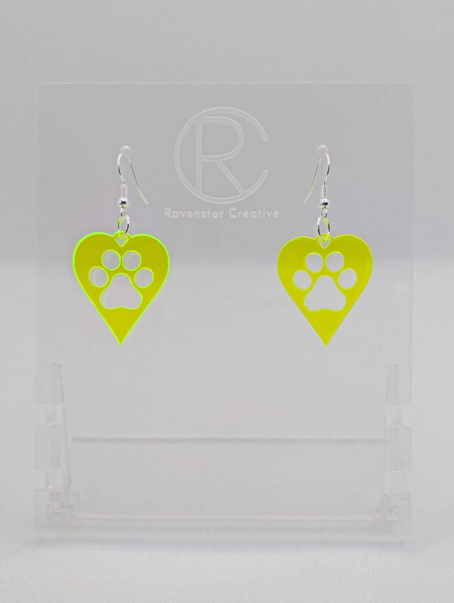 Shows a pair of earrings that have Silver ear wire hook fixings. They are hearts with a dog paw cut out of the middle, perfect for the puppy lover. The colour of them is Neon Green. They are on see through stands. Against a white background.