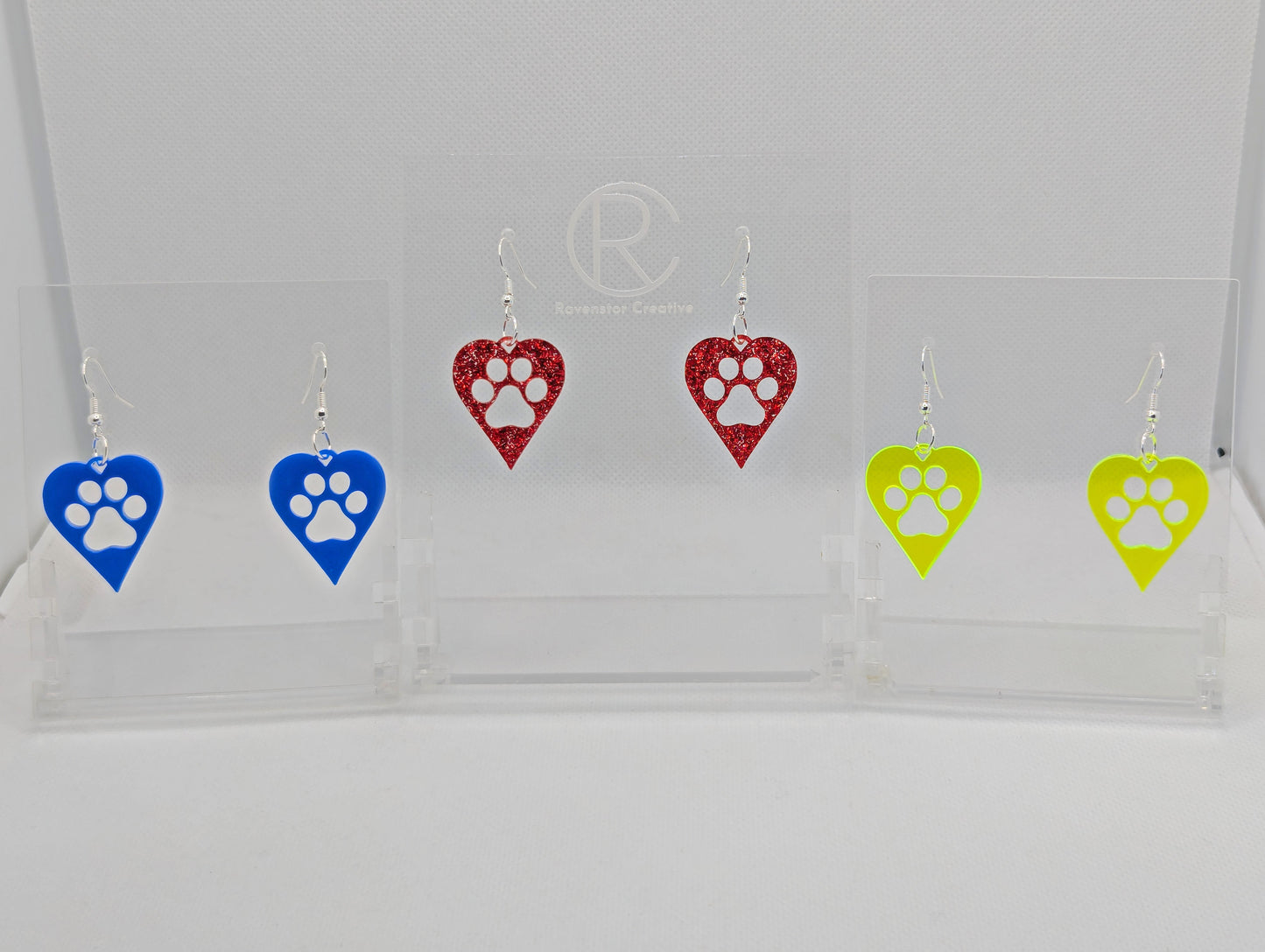 Shows three pairs of earrings that have ear wire hook fixings. They are hearts with a dog paw cut out of the middle, perfect for the puppy lover. The colours ate Blue, Red Glitter and Neon Green. They are on see through stands.