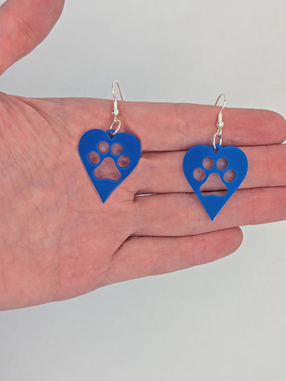 Shows a pair of earrings that have Silver ear wire hook fixings. They are hearts with a dog paw cut out of the middle, perfect for the puppy lover. They are against a hand to show the size of one and a half finger widths in length and same again wide